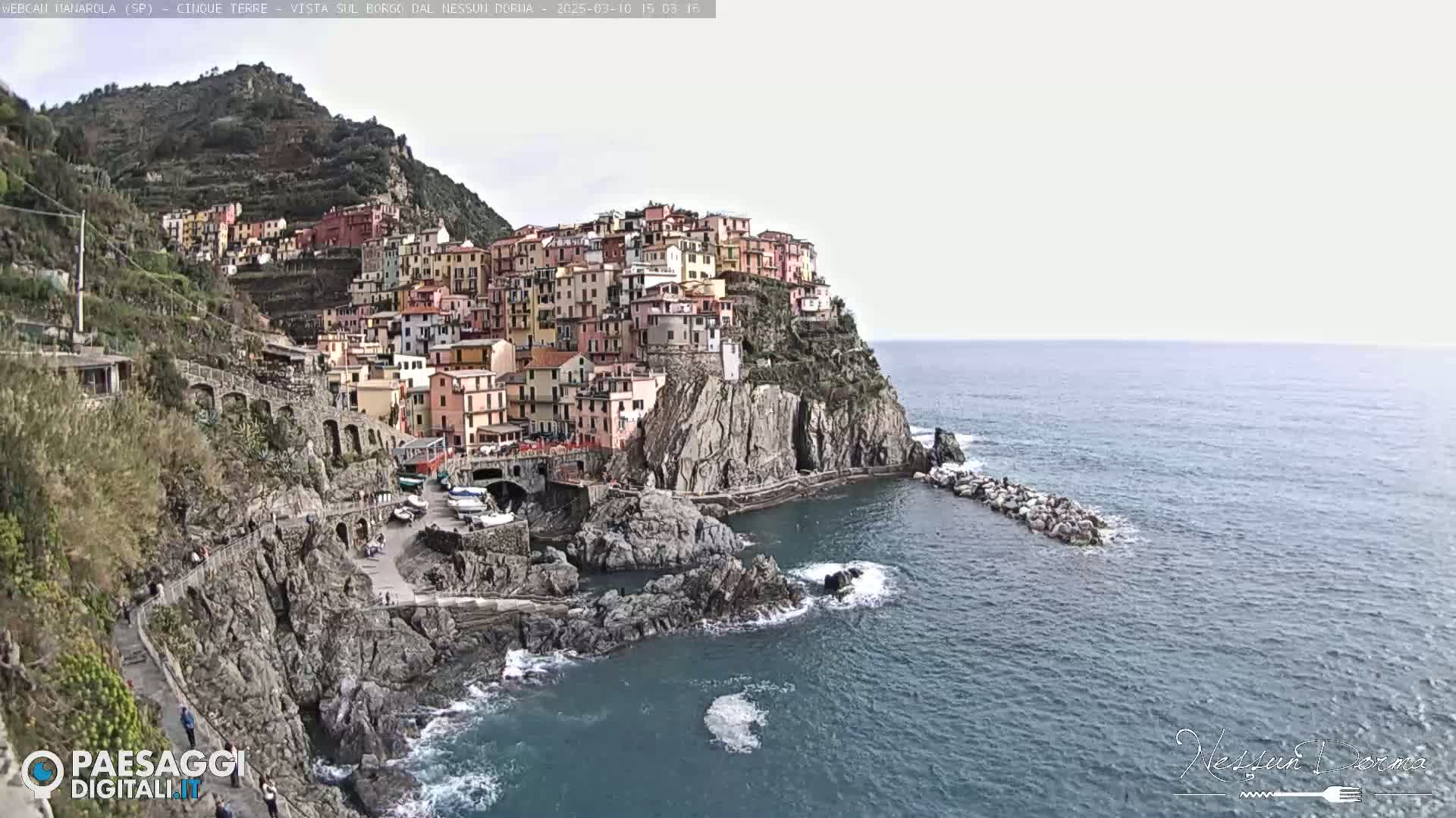 A colorful village clings to a steep cliffside overlooking a calm sea under a bright, overcast sky.