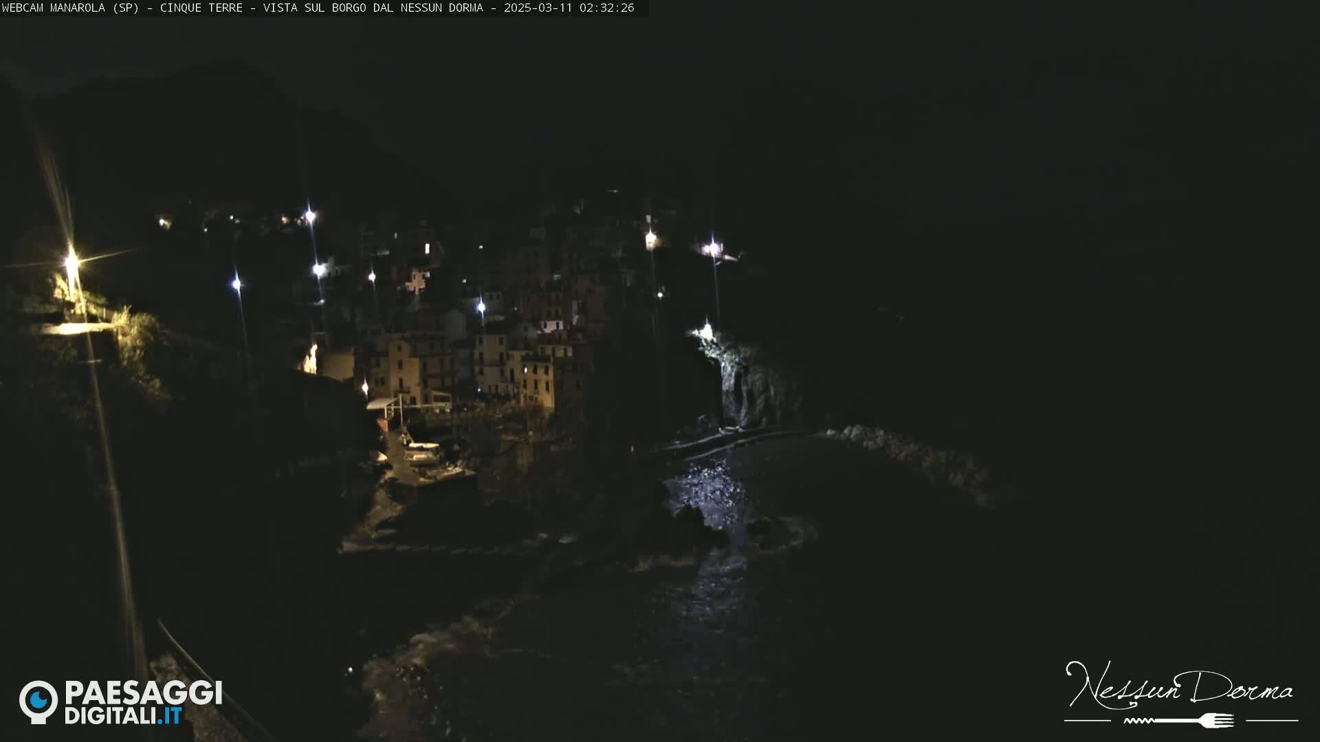 A nighttime, dimly lit coastal village clings to a dark hillside, with waves gently lapping at the shore below.