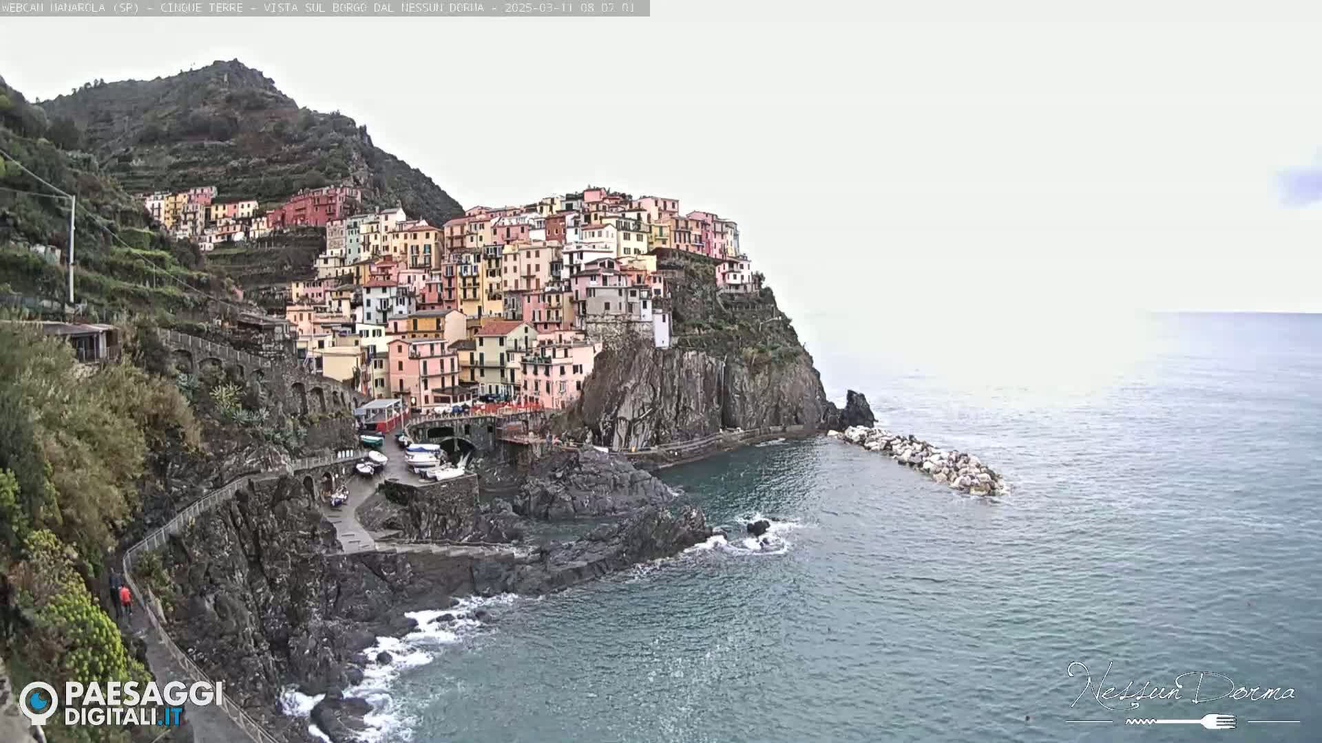 A colorful village clings to a steep cliffside overlooking a calm sea under an overcast sky.