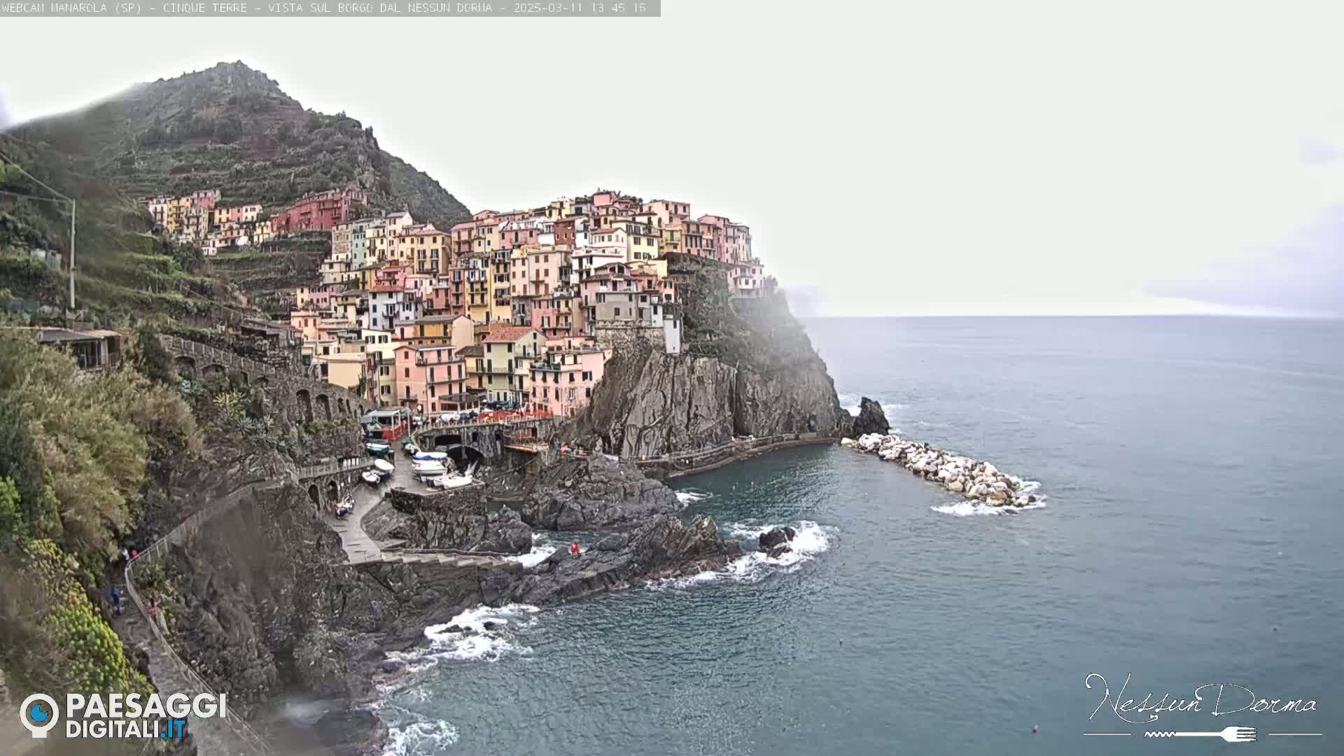 A colorful village clings to a steep cliffside overlooking a calm sea on an overcast day.