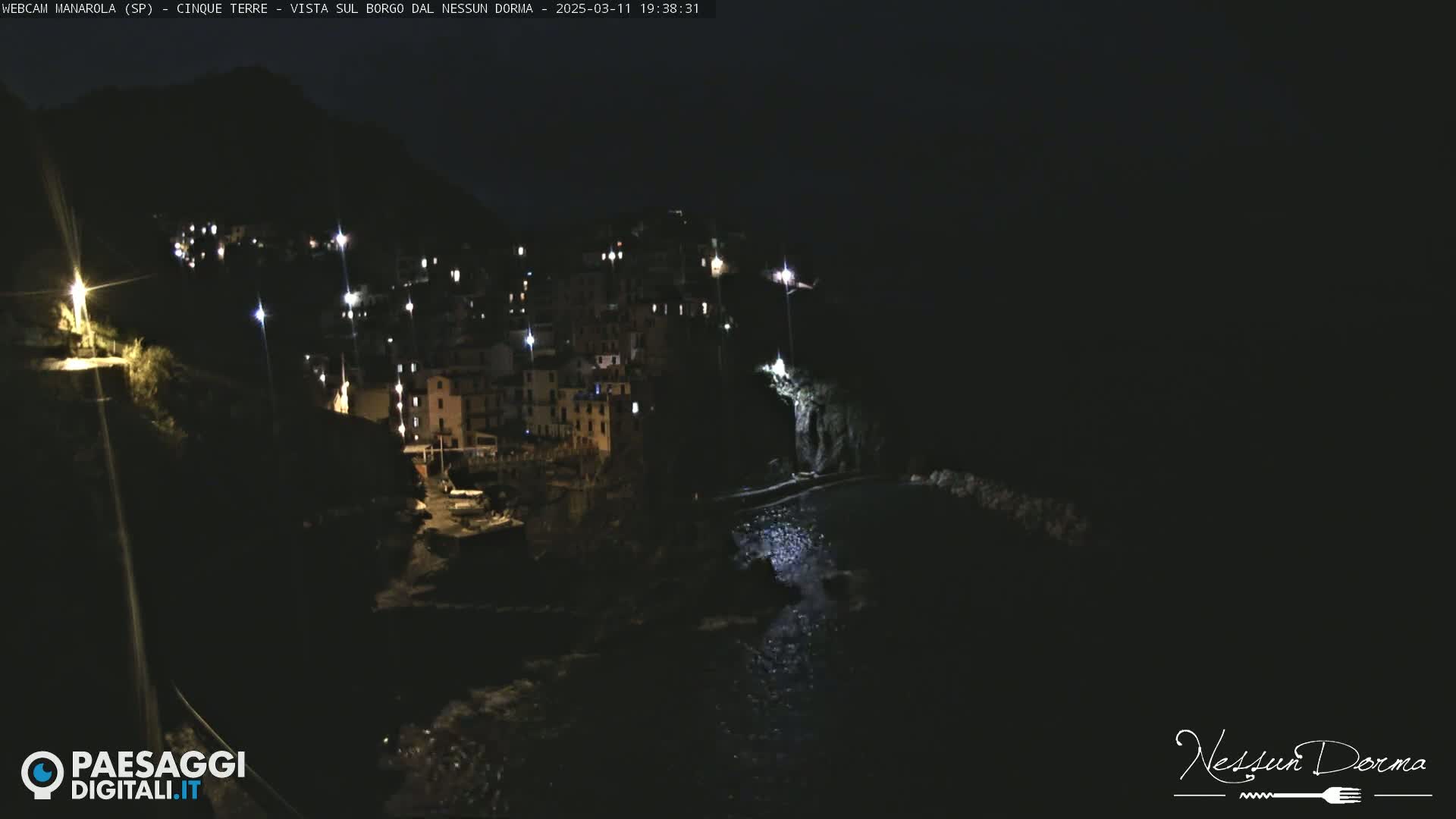 A nighttime view of a coastal village illuminated by lights, with dark water below.