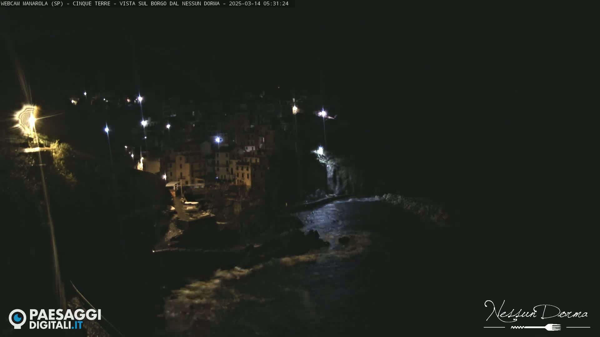 A nighttime view of a coastal town illuminated by lights, with dark water churning below.