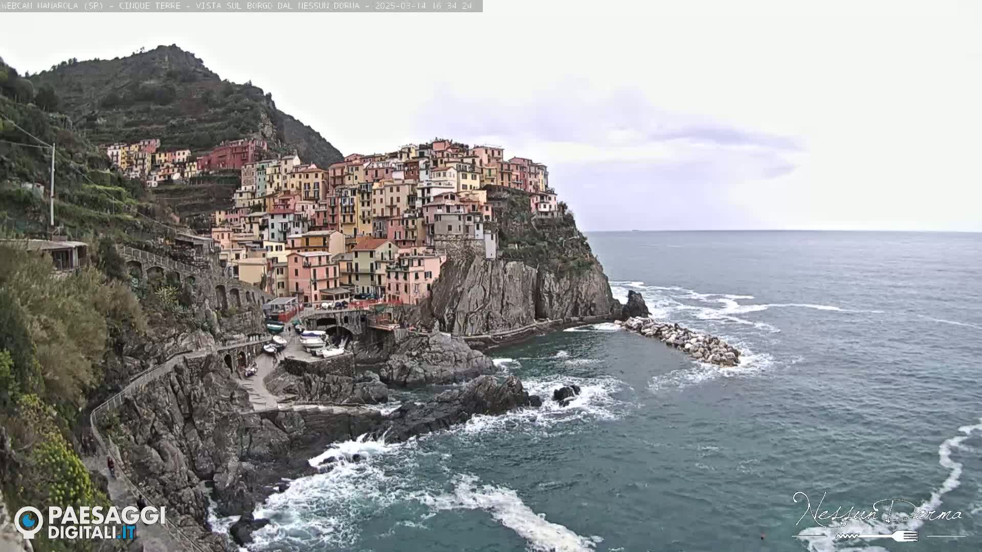 A colorful village clings to a rocky cliffside overlooking a choppy sea under an overcast sky.