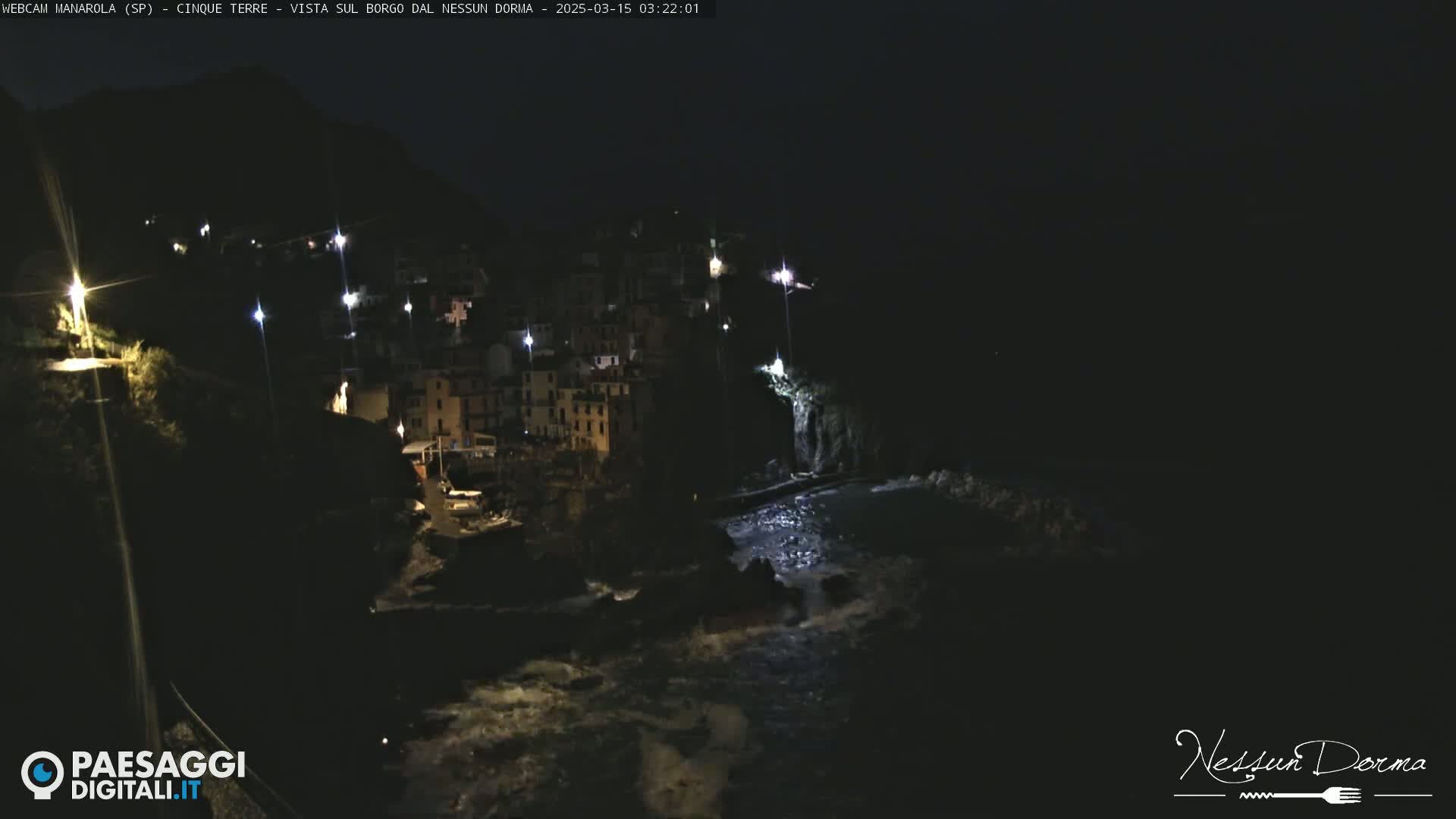 A nighttime coastal town with buildings illuminated, situated on a dark cliff overlooking a wavy sea.