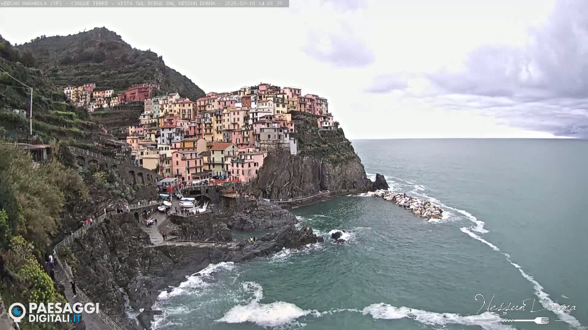 A colorful village clings to a steep cliffside overlooking a choppy sea under a cloudy sky.