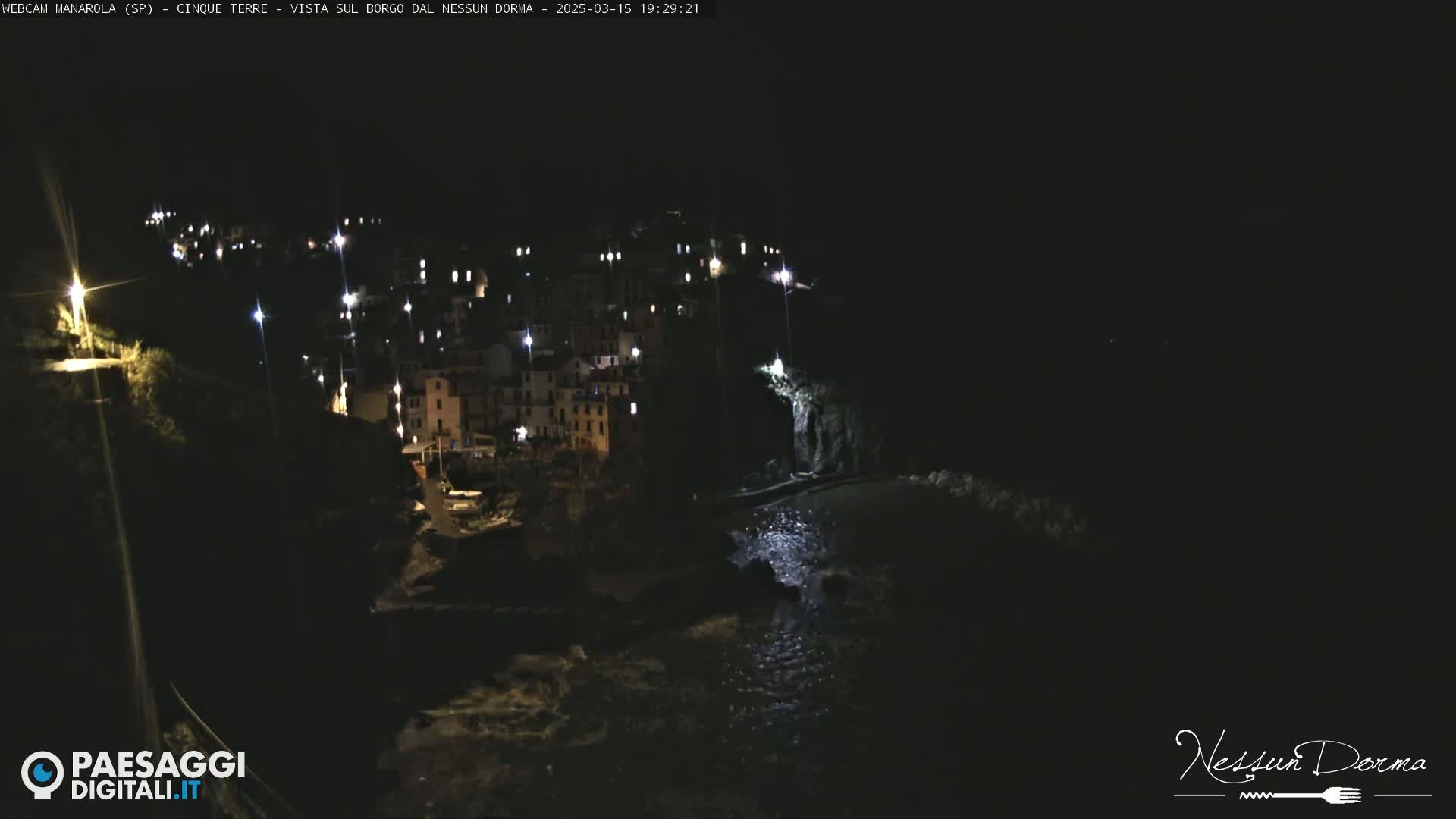 A nighttime, low-angle view of a coastal village illuminated by lights, with dark water lapping at the shore.