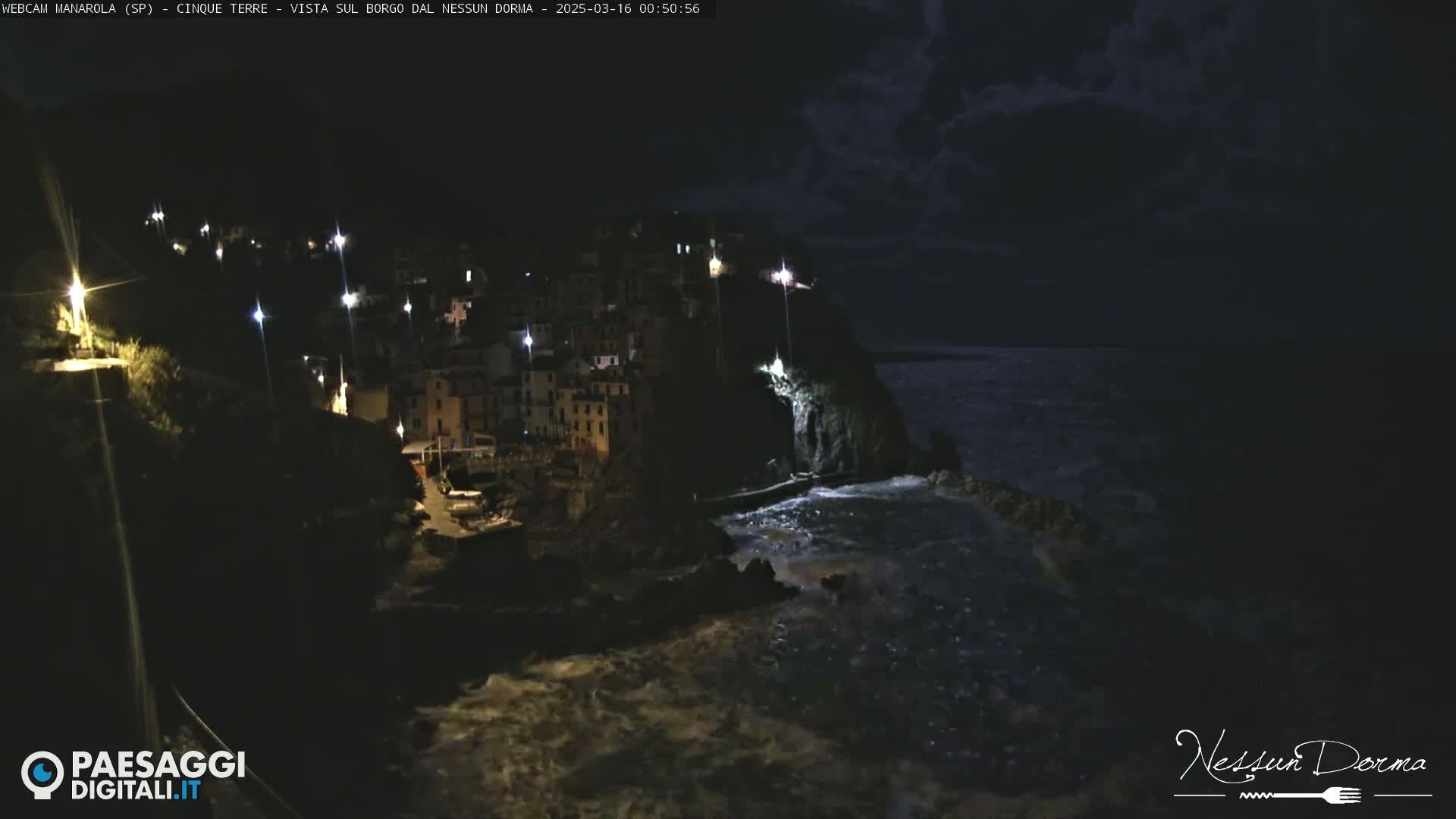 A nighttime coastal village with illuminated buildings is perched on a cliff overlooking a dark, slightly turbulent sea under a cloudy sky.