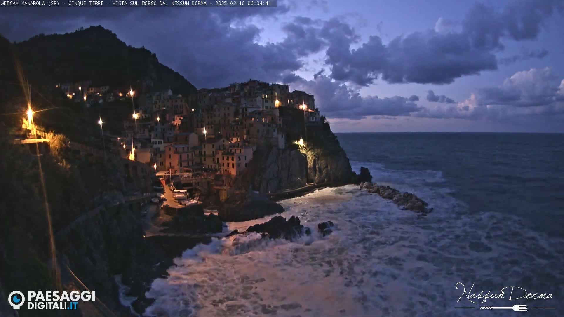 A village of lights clinging to a cliffside overlooking a dark, turbulent sea under a cloudy twilight sky.