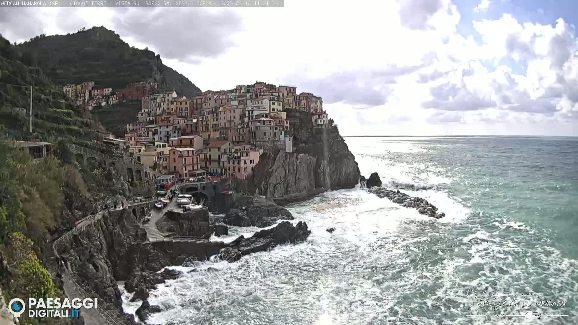 A colorful village clings to a steep cliffside overlooking a turbulent sea under a partly cloudy sky.