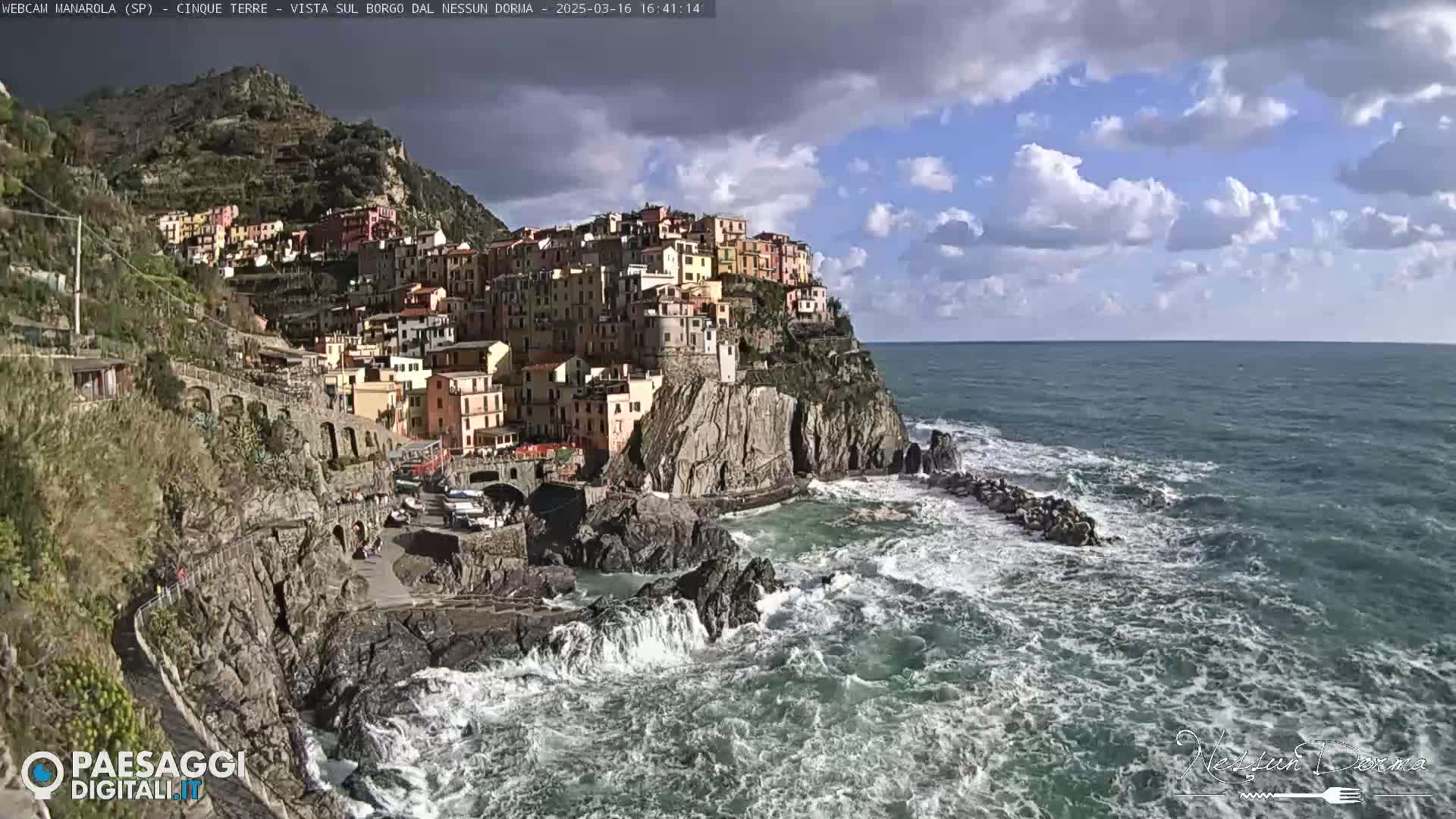 A colorful village clings to a steep cliffside overlooking a turbulent sea under a partly cloudy sky.