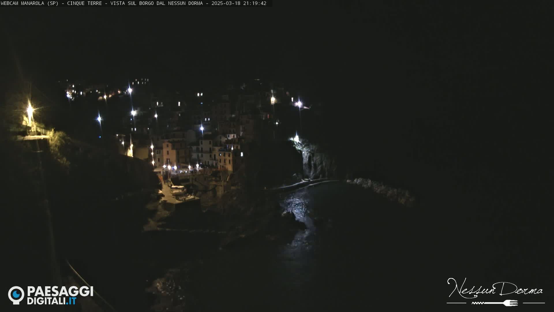 A nighttime, aerial view of a small, coastal town nestled against a dark cliffside, with its lights illuminated against the black water.
