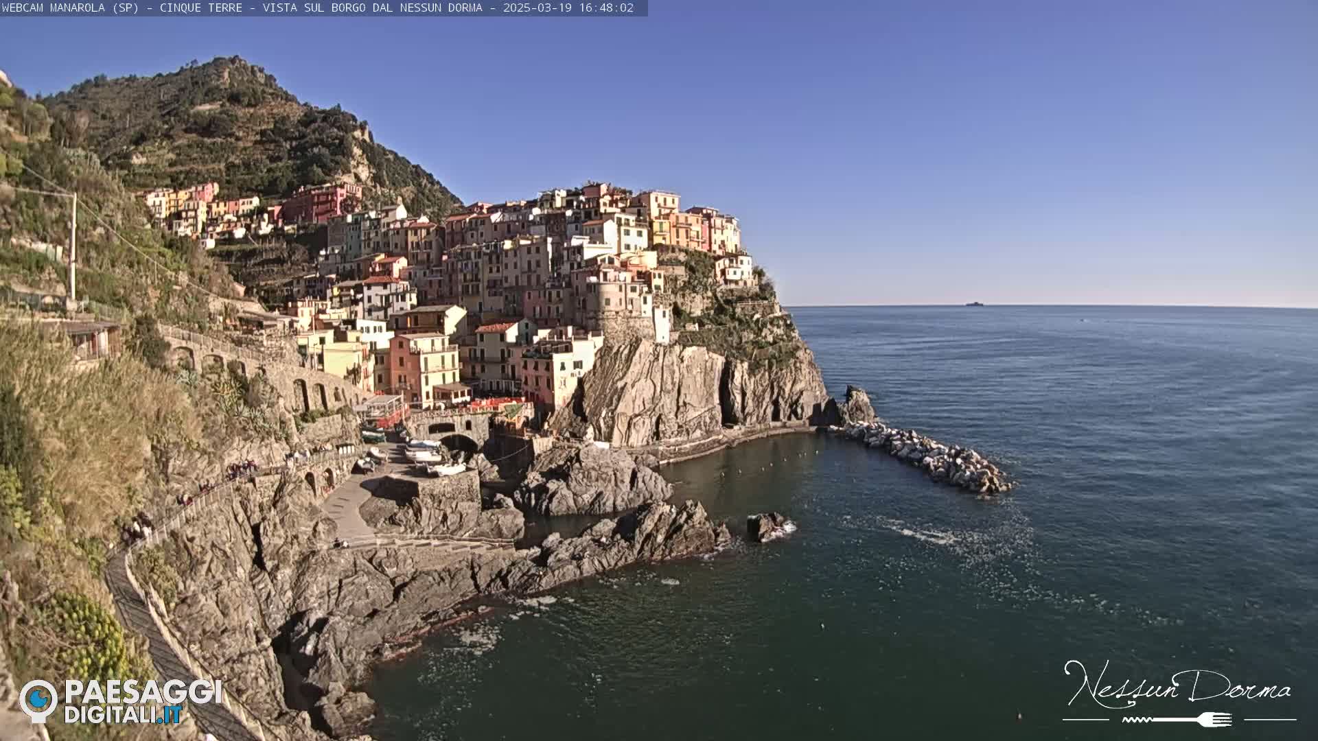 A colorful village clings to a rocky cliffside overlooking a calm, blue sea under a clear sky.