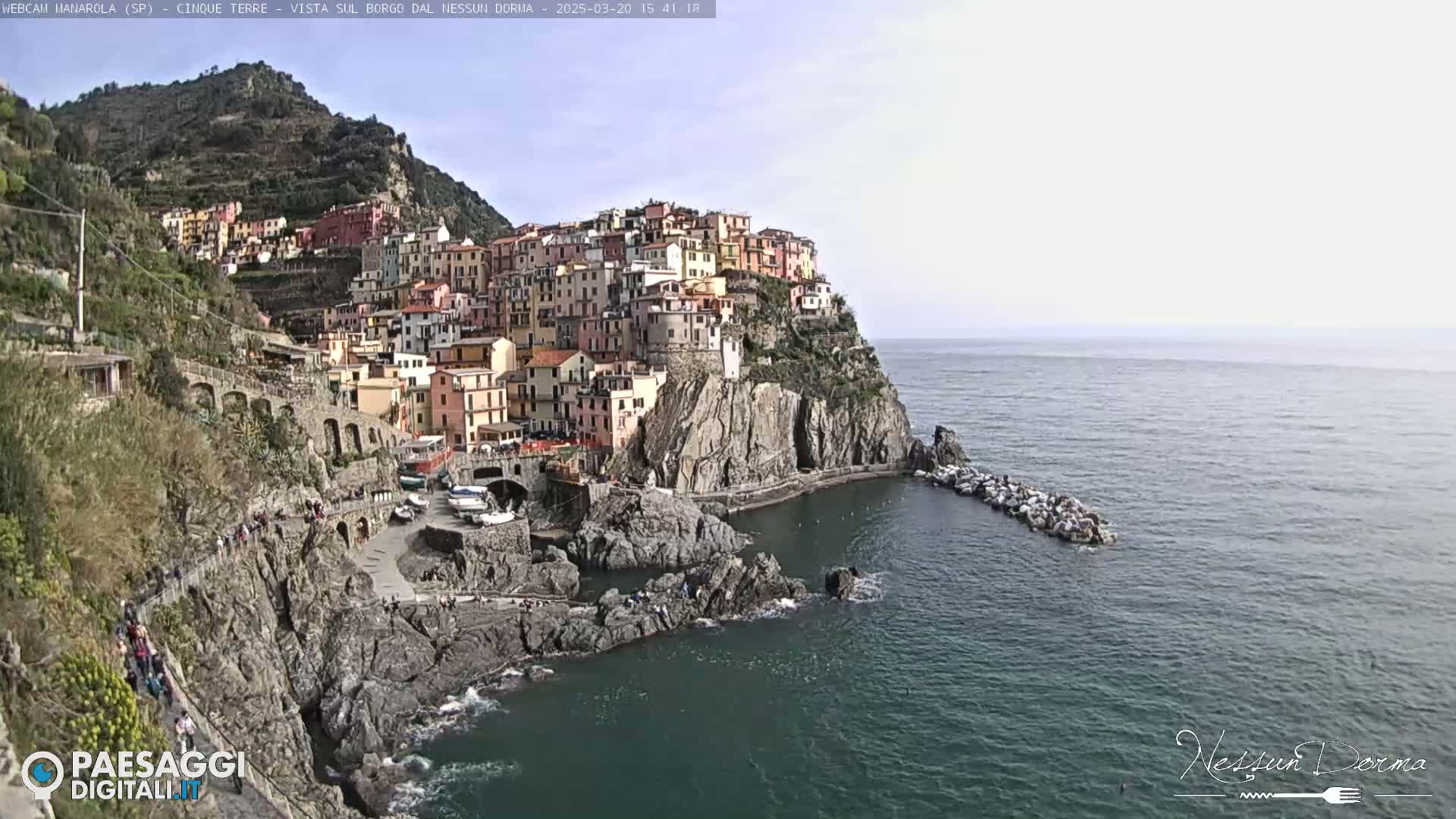 A colorful village clings to a steep cliffside overlooking a calm sea under a mostly clear sky.