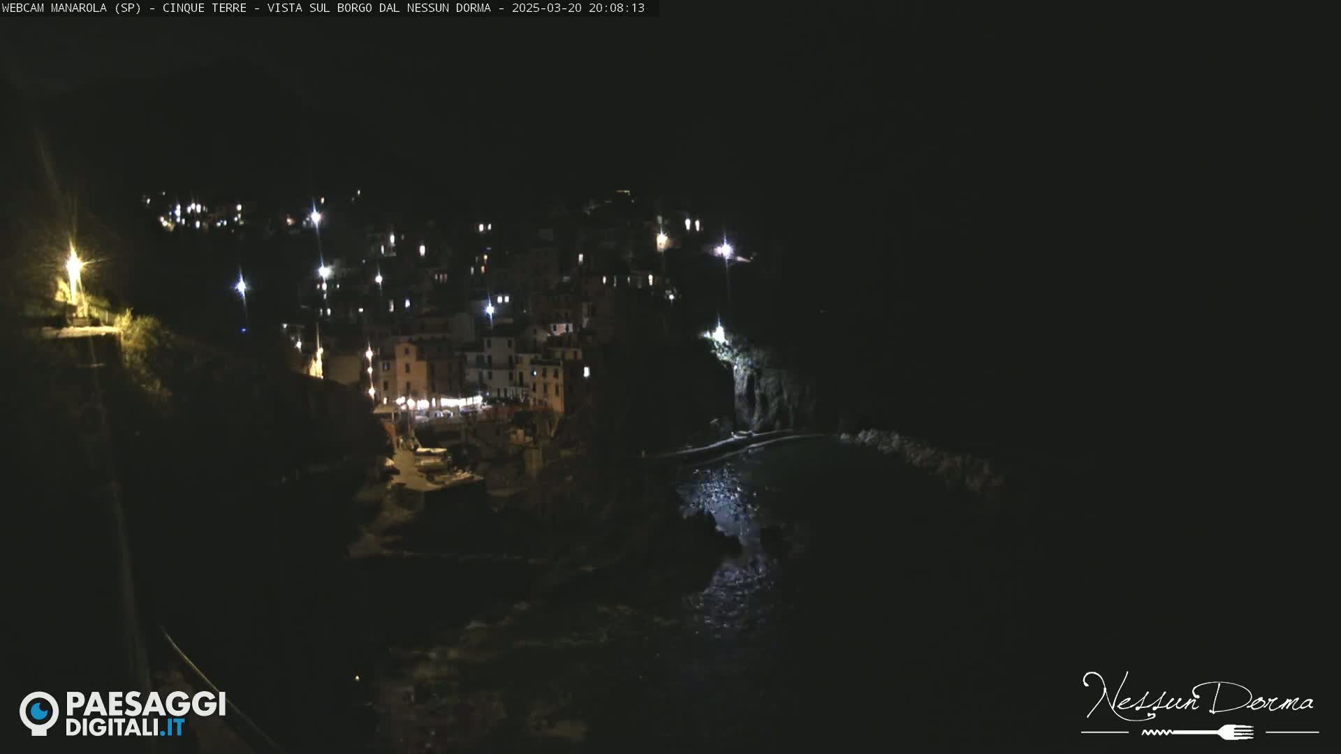 A nighttime, coastal town with lights illuminating buildings clinging to a dark hillside, partially visible over dark water.