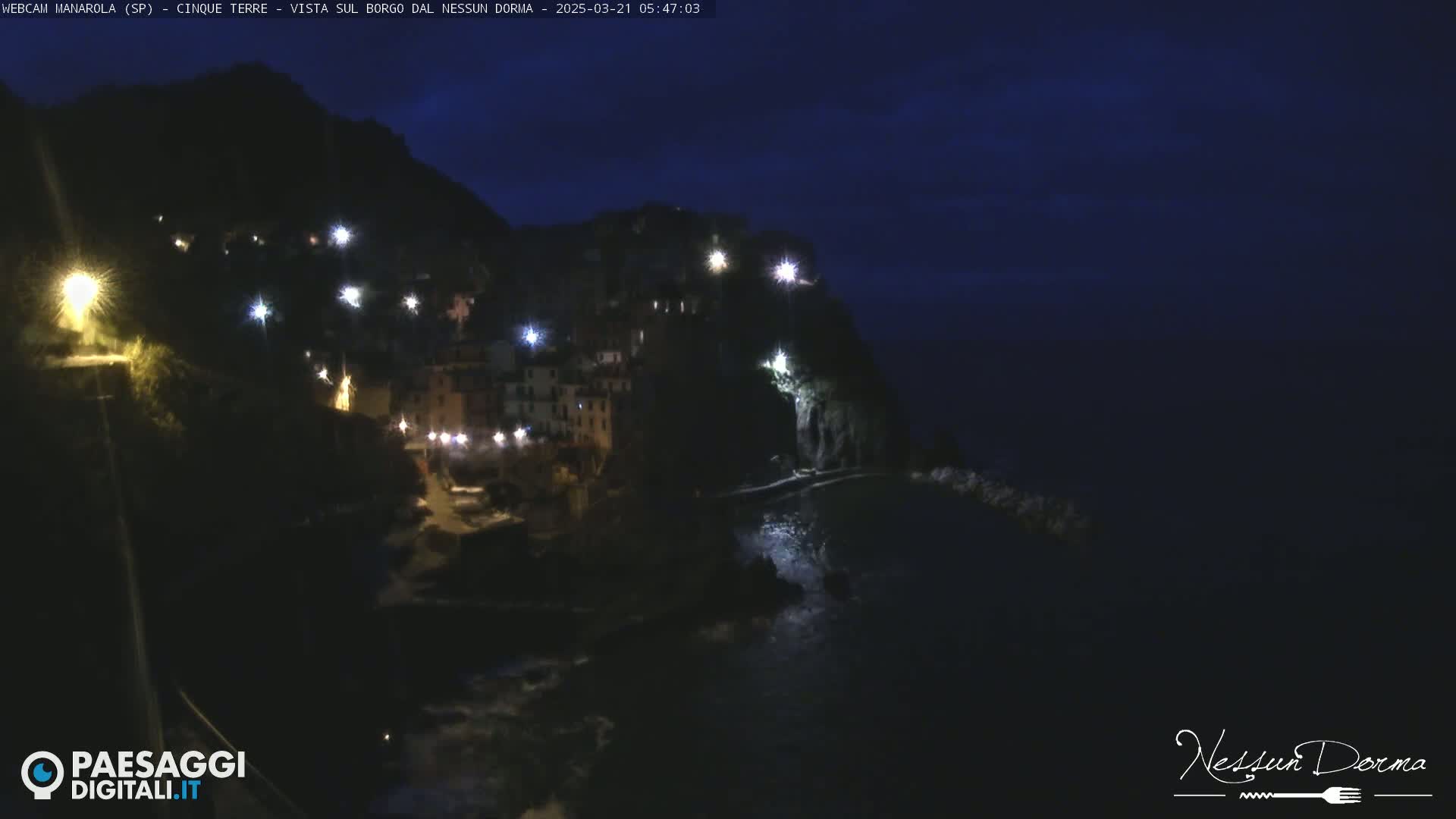 A dark, nighttime view of a coastal town nestled on a cliffside, illuminated by scattered lights, with a dark ocean below.