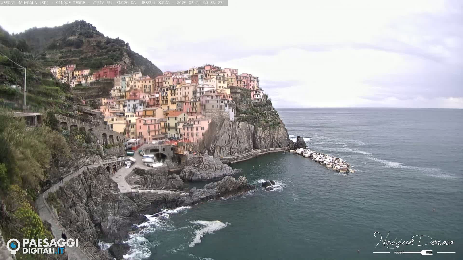 A colorful village clings to a steep cliffside overlooking a calm, gray sea under an overcast sky.