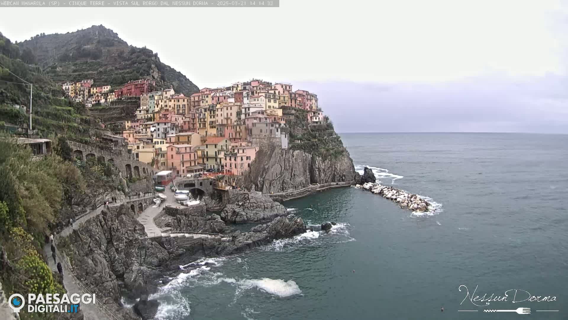 A colorful village clings to a steep cliffside overlooking a calm, gray sea under an overcast sky.