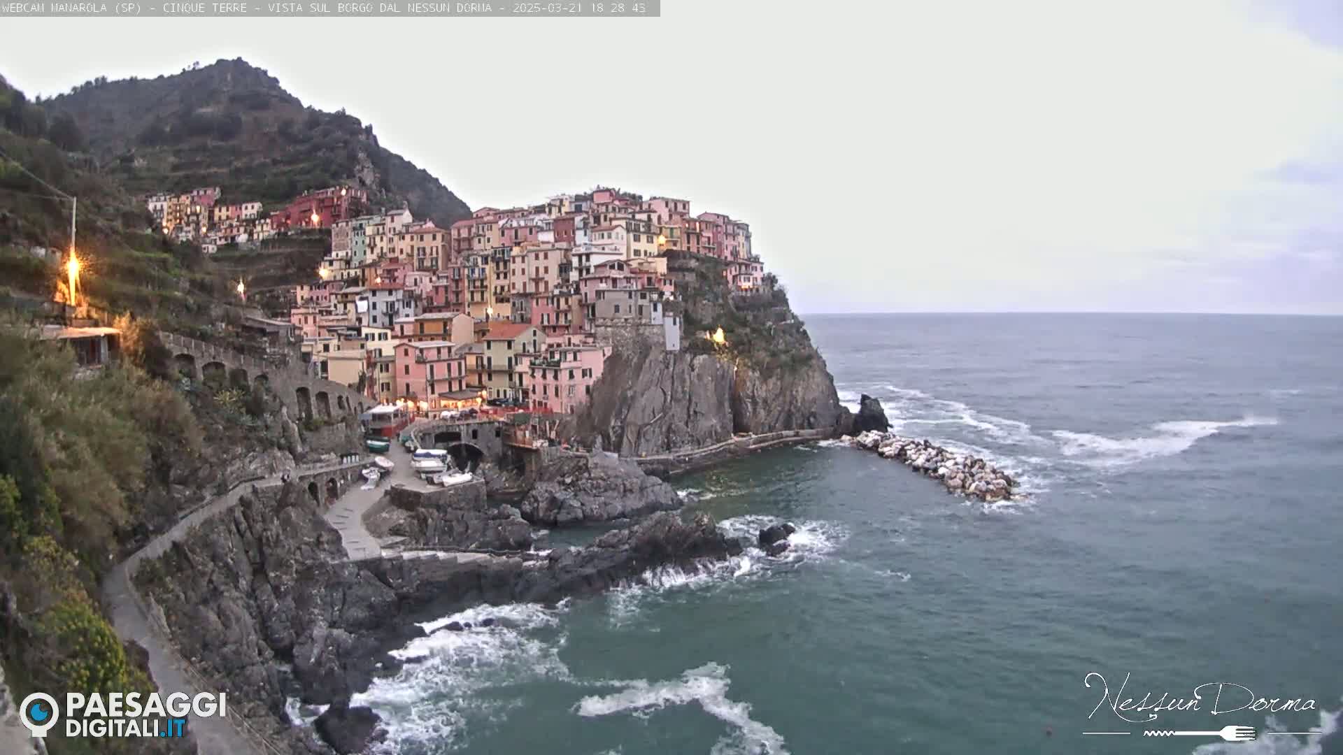A colorful village clings to a steep cliffside overlooking a calm, gray sea under an overcast sky.