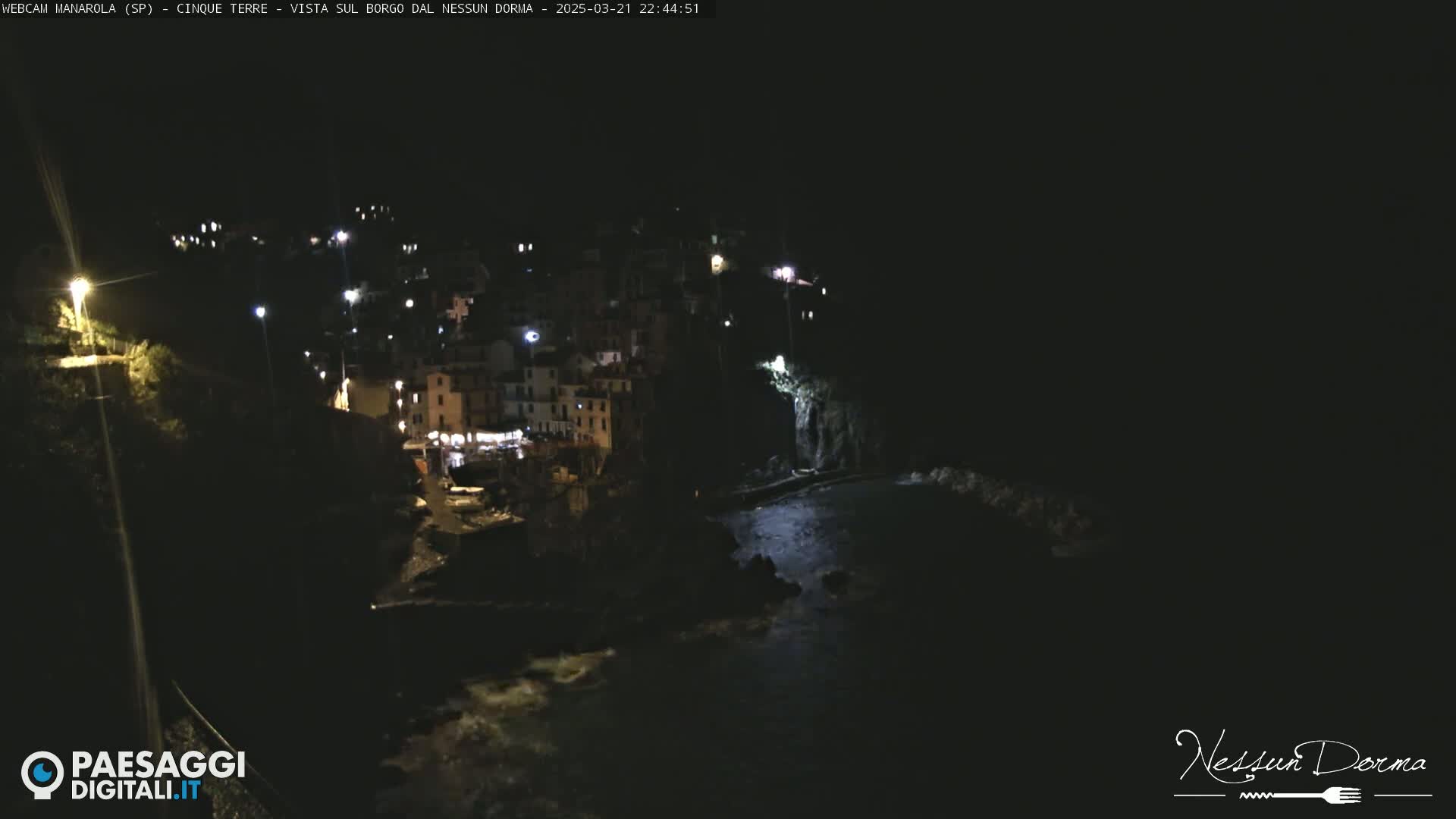 A nighttime, aerial view shows a small, coastal town illuminated by scattered lights, nestled against dark cliffs and a body of water.