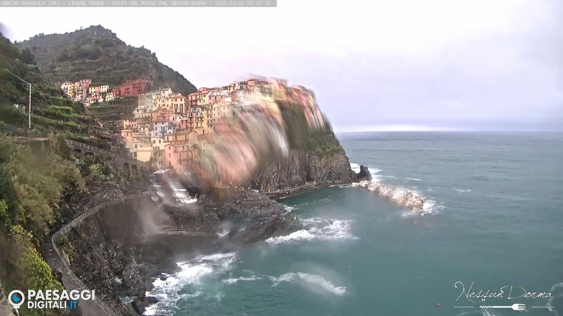 A colorful village clings to a steep cliffside overlooking a turbulent sea under an overcast sky.