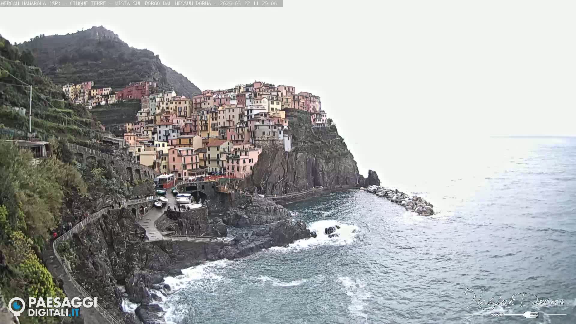 A colorful village clings to a steep, rocky cliffside overlooking a choppy sea under an overcast sky.