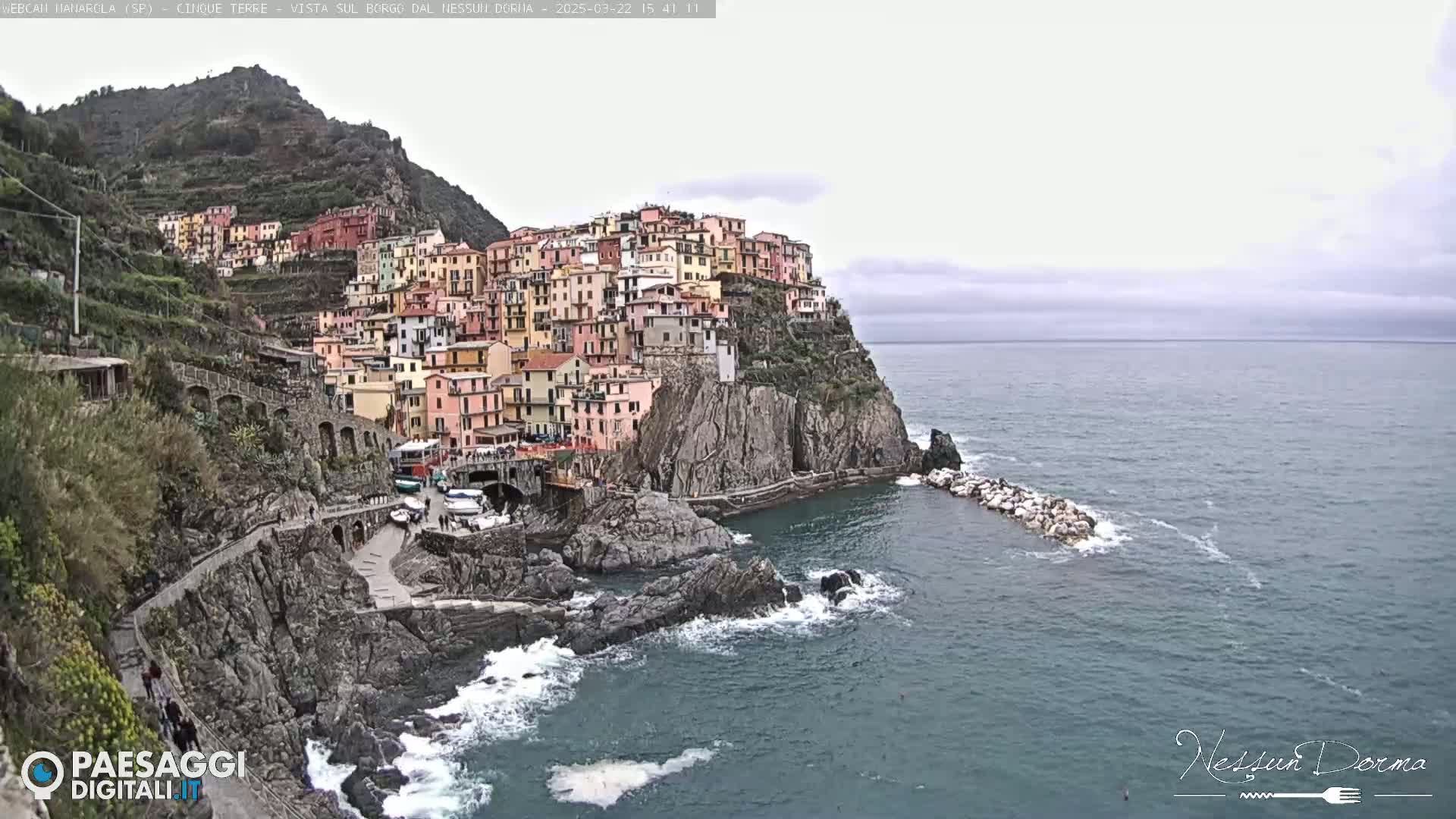 A colorful village clings to a steep cliffside overlooking a dark-blue sea under an overcast sky.