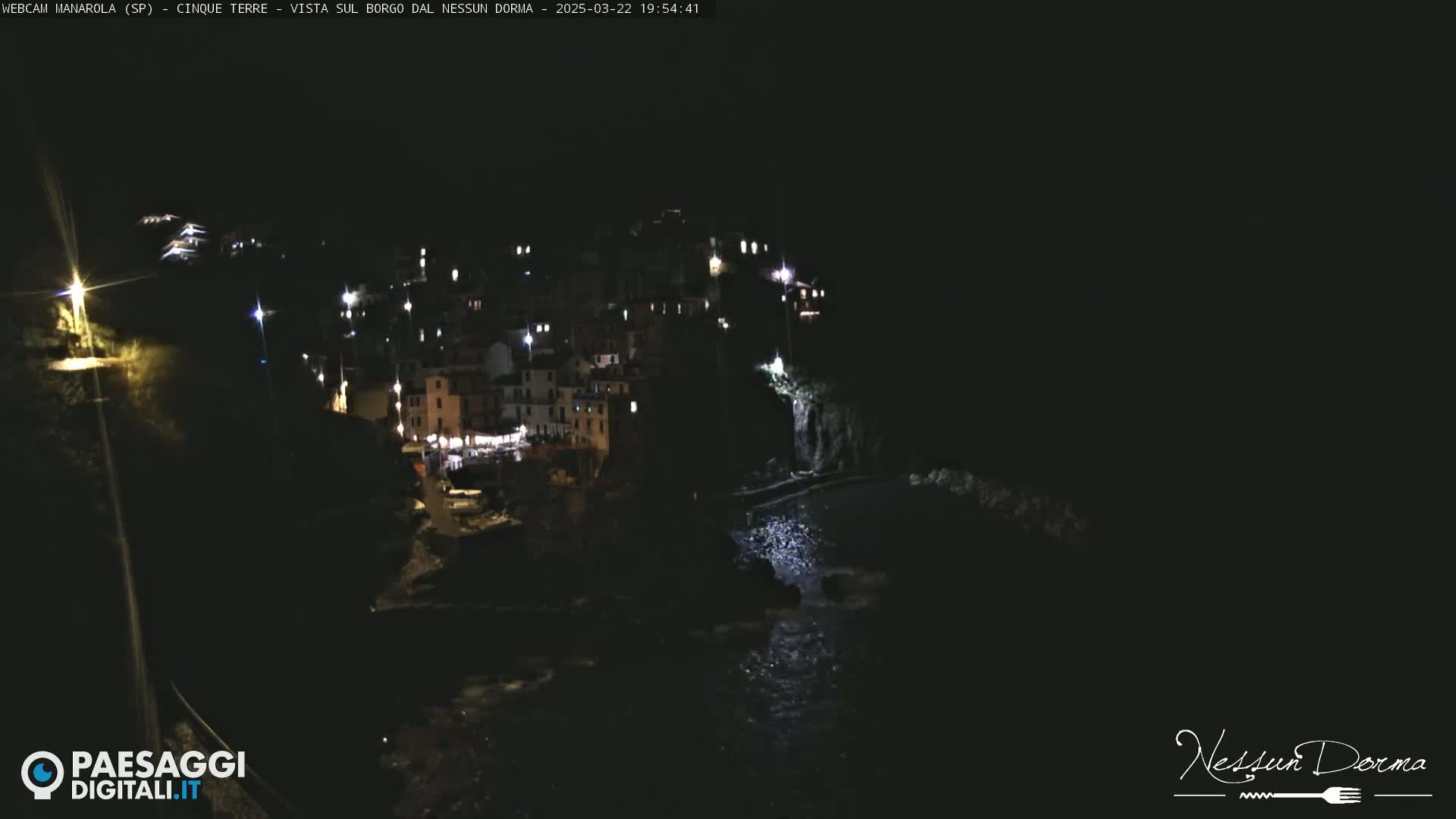 A nighttime view of a small, brightly lit coastal town nestled on a dark cliffside overlooking the water.