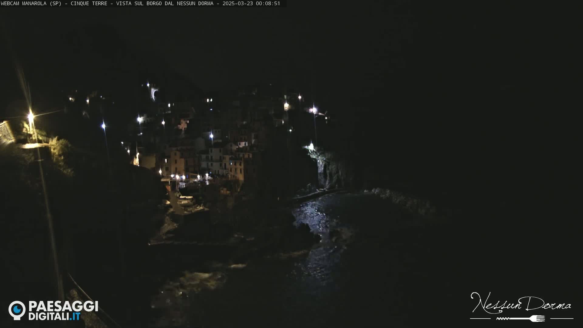 A nighttime view of a coastal town, illuminated by lights, with dark water below.