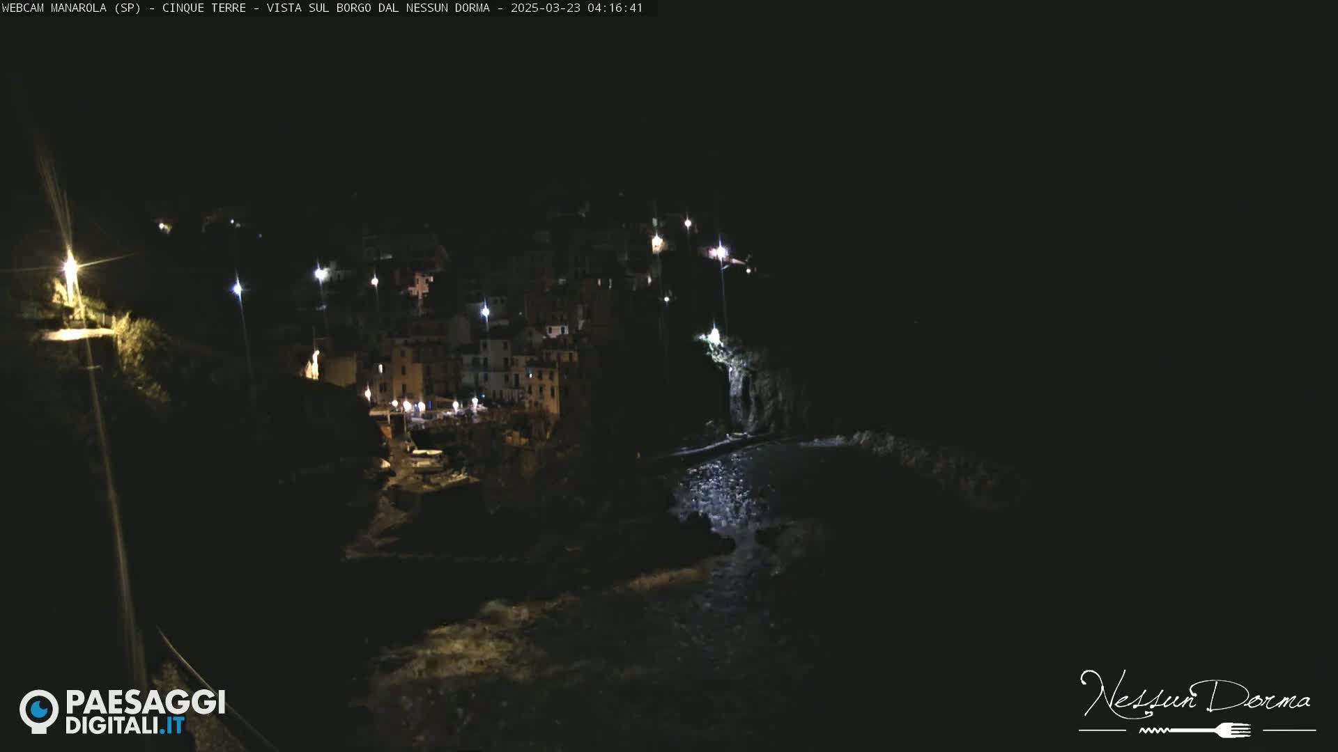 A nighttime view of a coastal town illuminated by streetlights, with dark water flowing in the foreground.