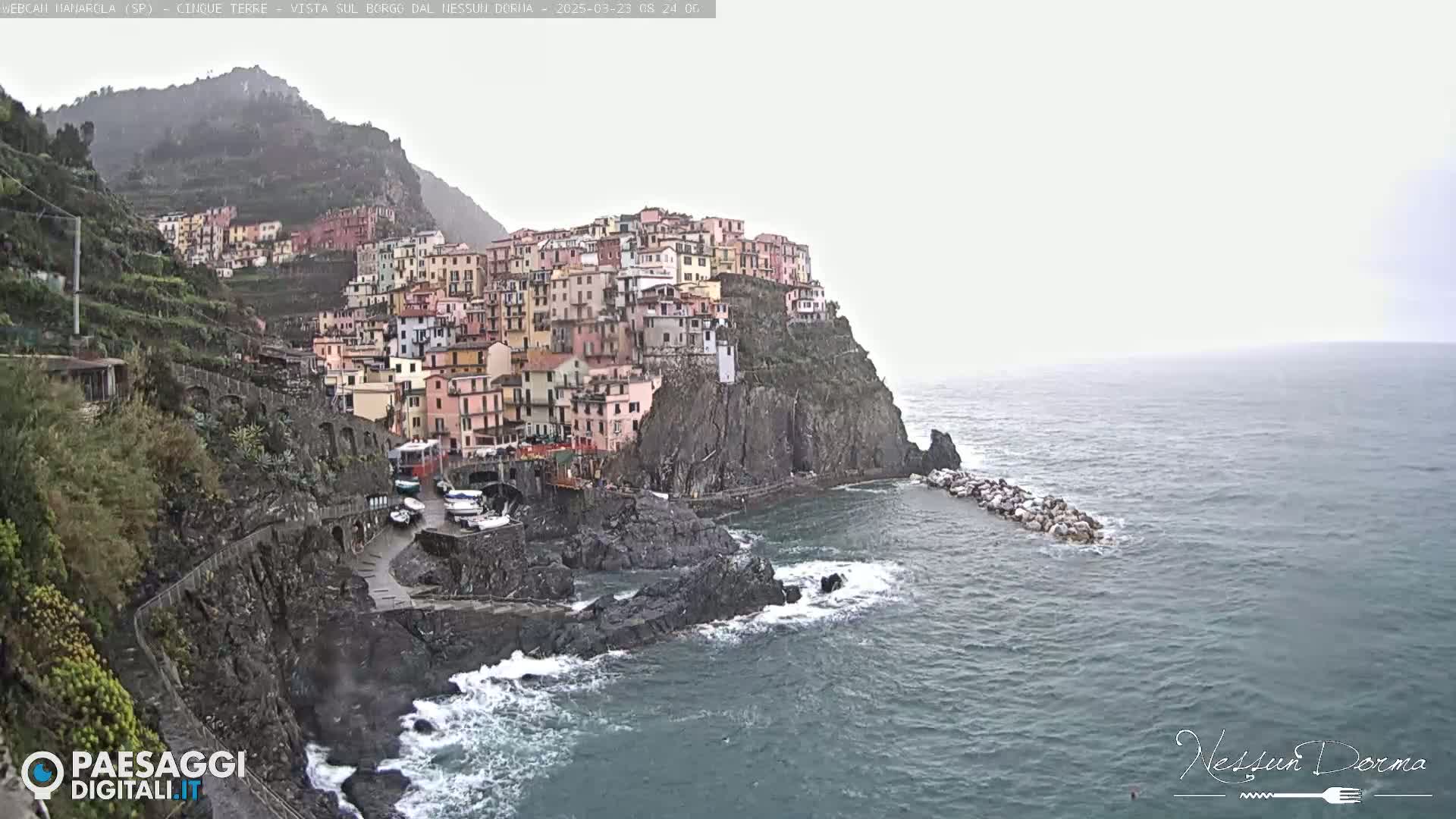 A colorful village clings to a steep cliffside overlooking a rough sea under an overcast sky.