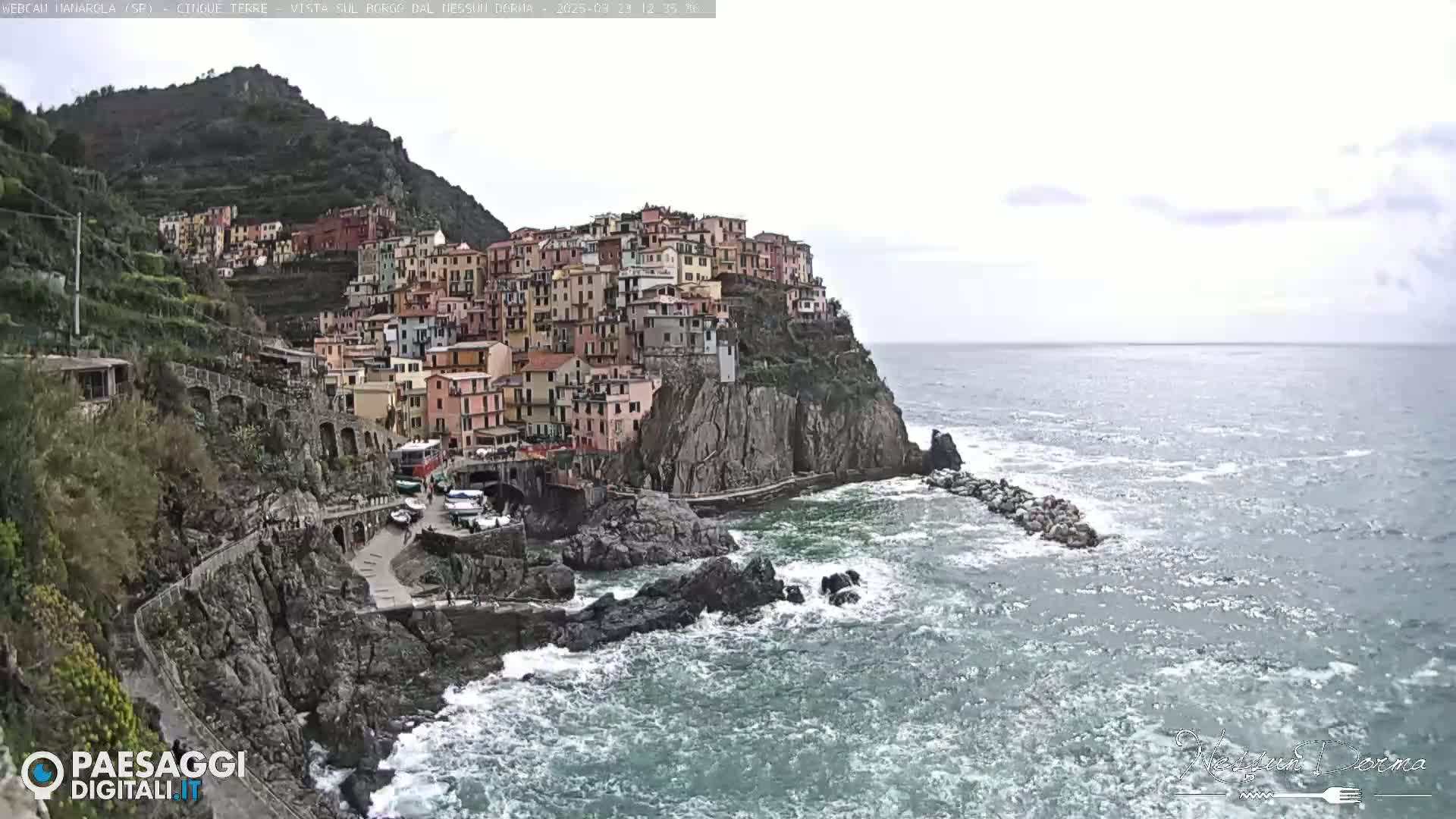 A colorful village clings to a steep cliffside overlooking a choppy sea under an overcast sky.