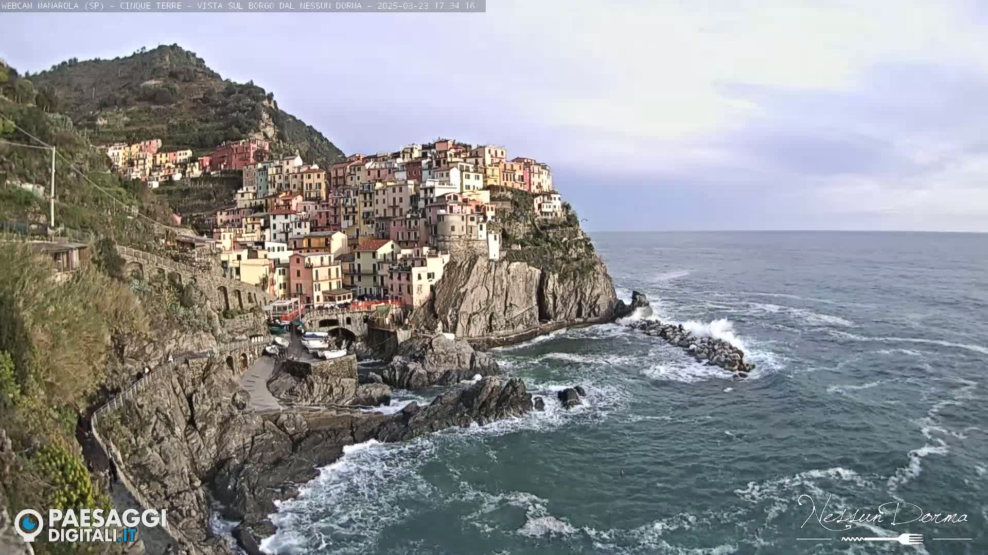 A colorful village clings to a rocky cliffside overlooking a turbulent sea under a mostly cloudy sky.
