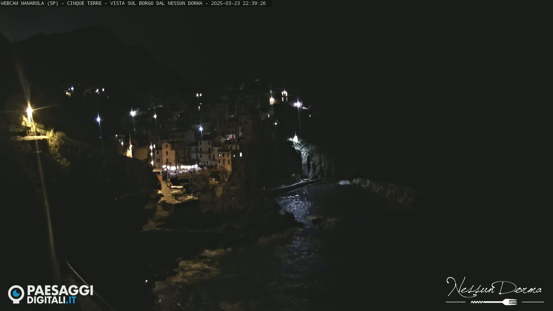 A nighttime, coastal village with buildings illuminated by lights is nestled against dark cliffs and a dark body of water.
