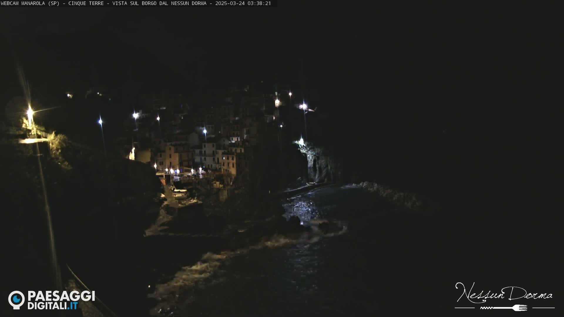 A nighttime, low-angle view of a coastal town illuminated by lights, with dark water below.