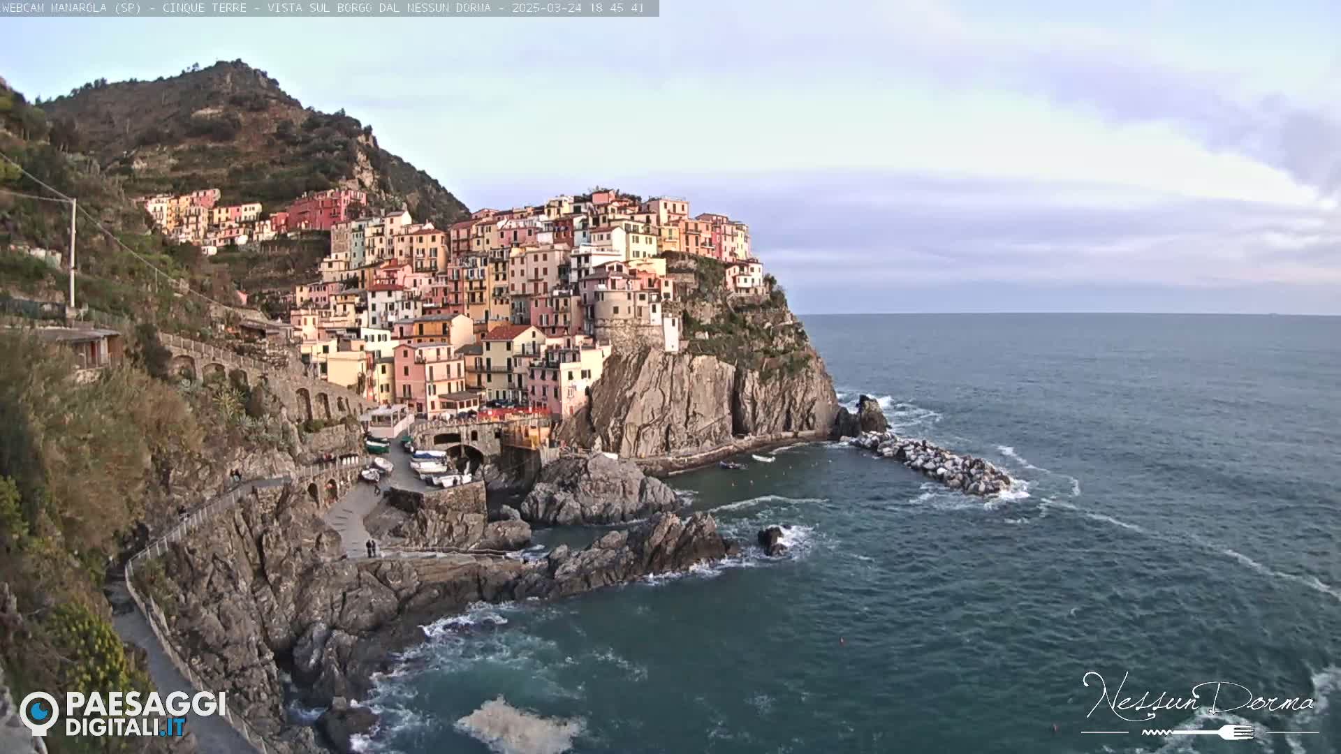 A colorful village clings to a rocky cliffside overlooking a calm sea under a mostly cloudy sky.