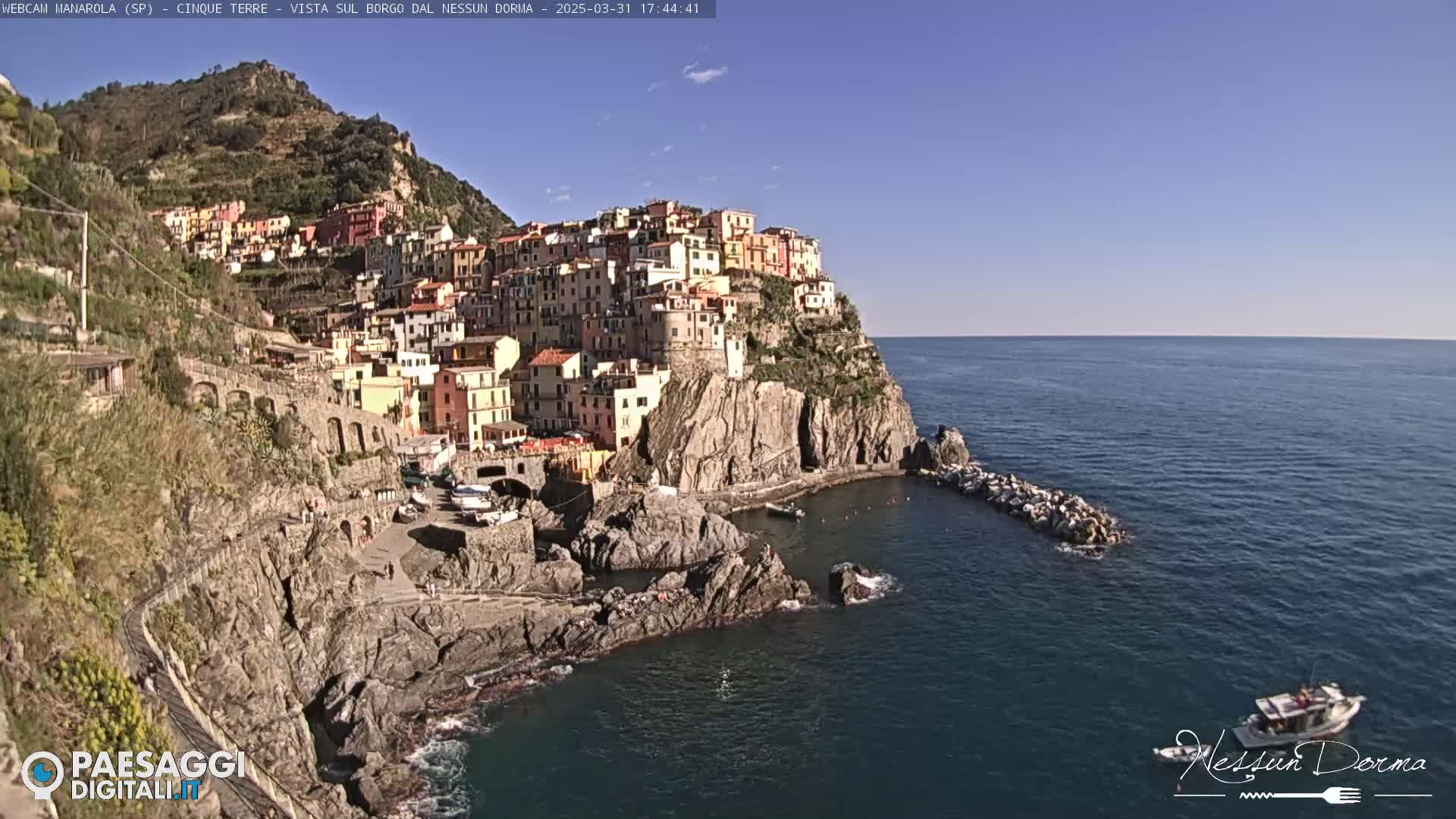 A colorful village clings to a rocky cliffside overlooking a calm blue sea under a clear blue sky.