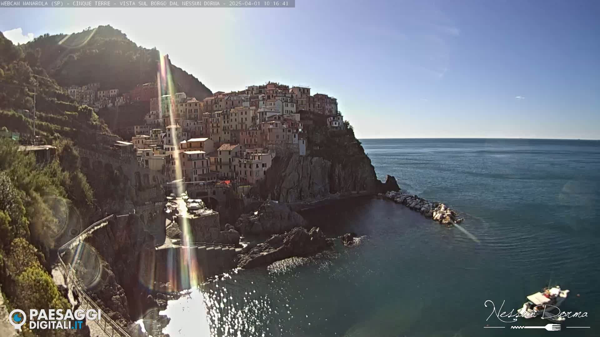 A colorful village clings to a rocky cliffside overlooking a calm, blue sea on a sunny day.