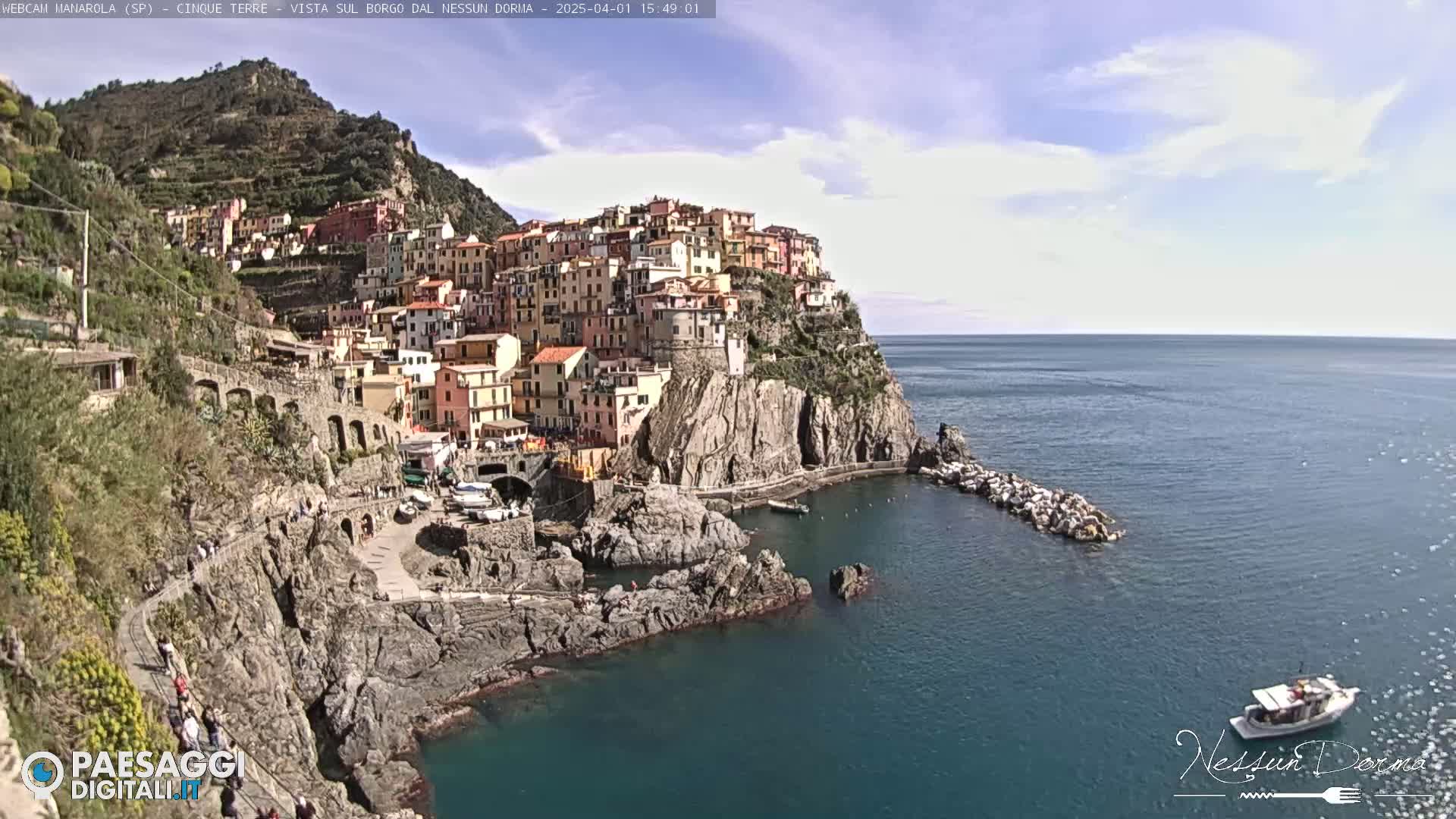 A colorful village clings to a rocky cliffside overlooking a calm, blue sea under a mostly sunny sky.