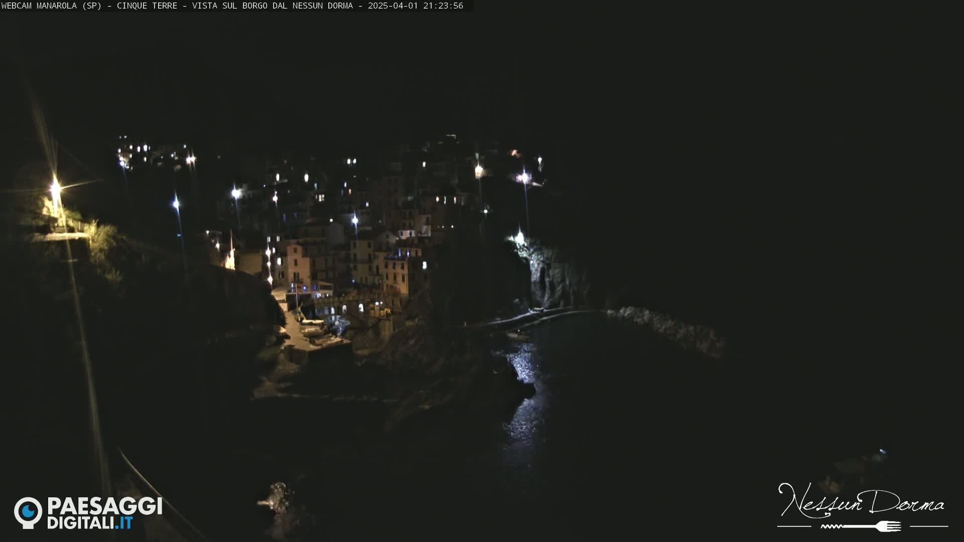 A nighttime view of a coastal town illuminated by lights, nestled between dark cliffs and a calm body of water.