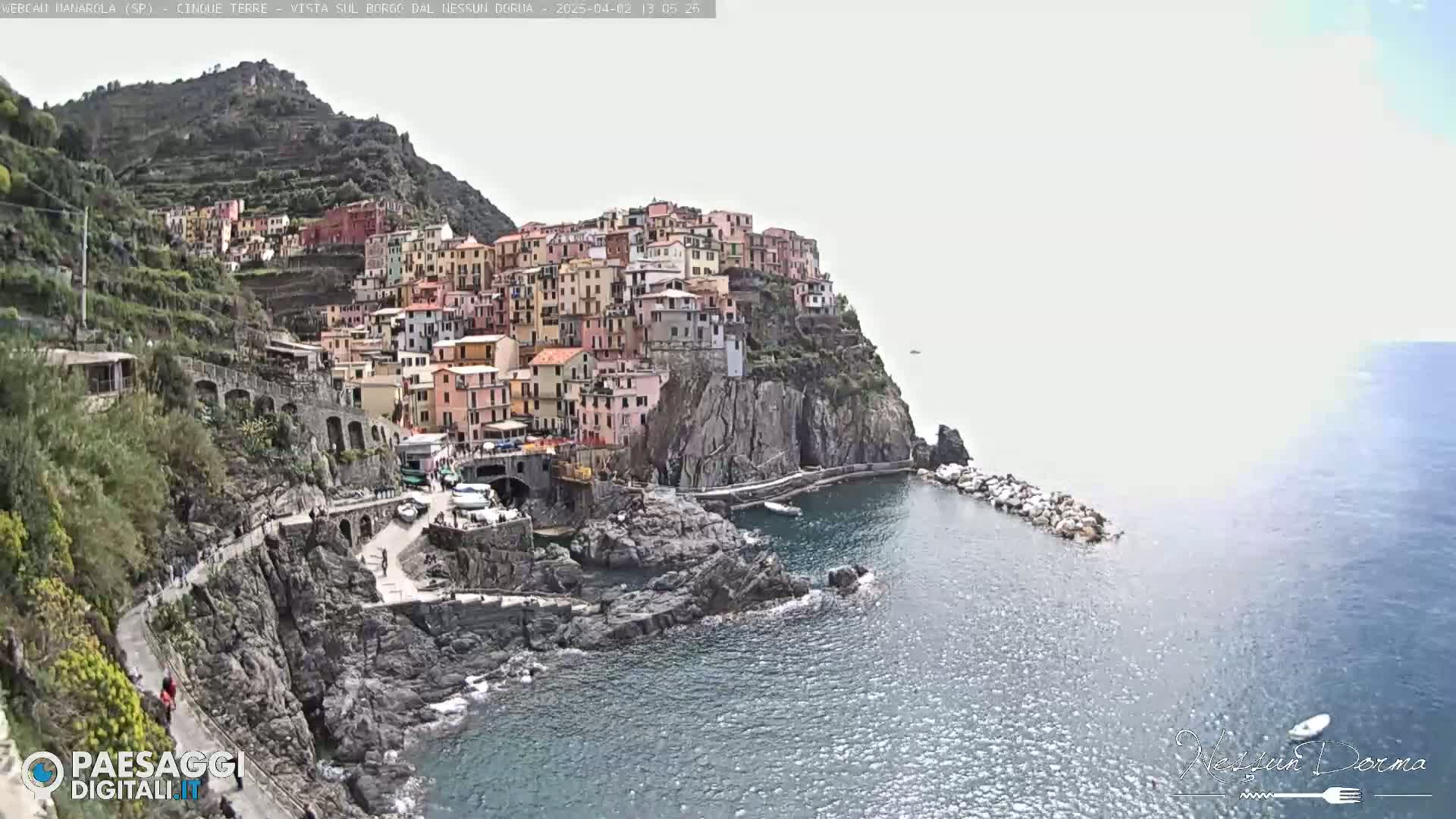 A colorful village clings to a steep, rocky hillside overlooking a calm sea.