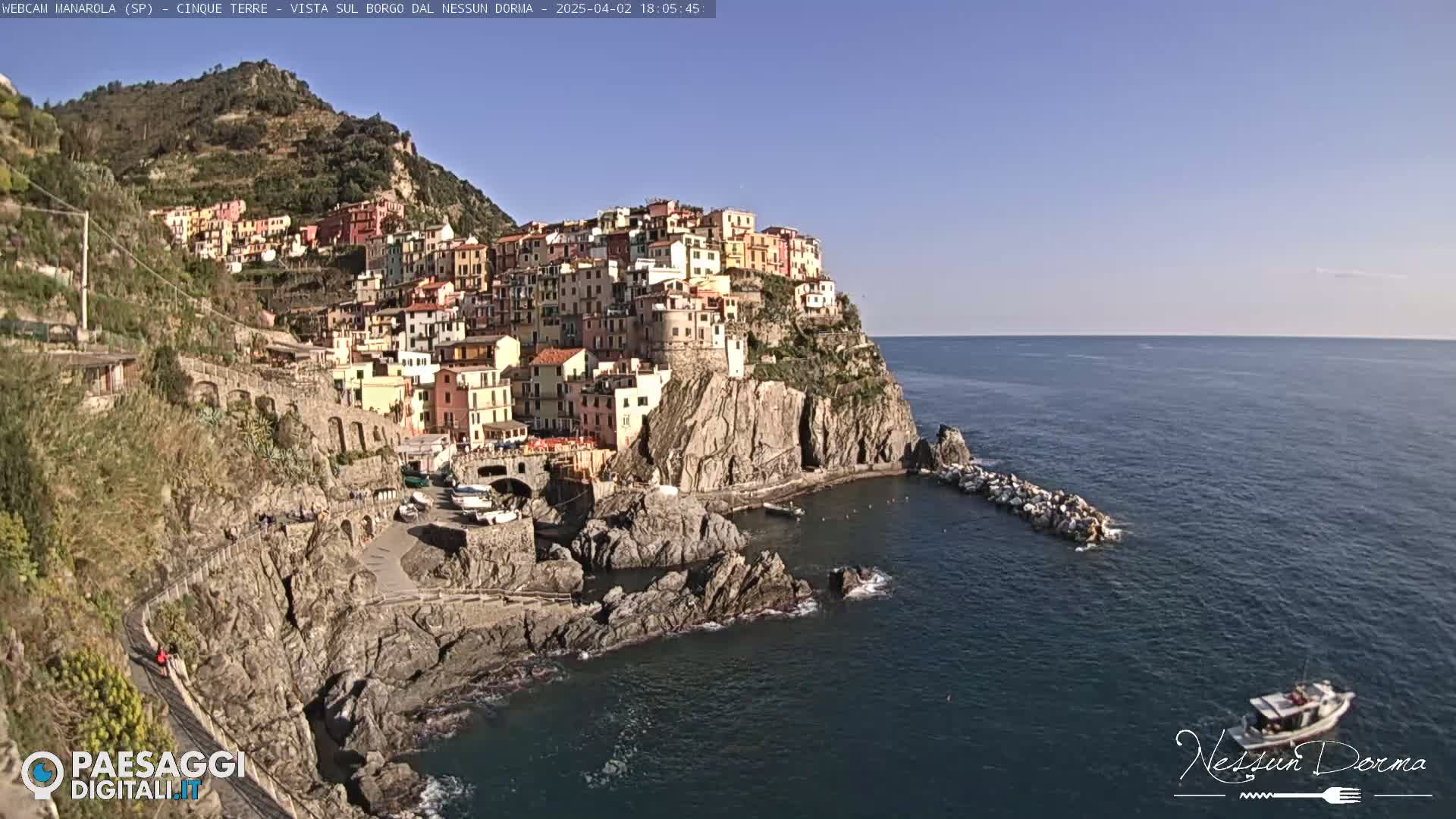 A colorful village clings to a rocky cliffside overlooking a calm, dark-blue sea under a clear, bright sky.