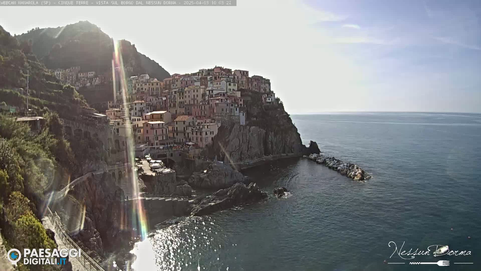 A brightly lit coastal town, built on a steep cliffside, overlooks a calm, dark blue sea under a mostly clear sky.