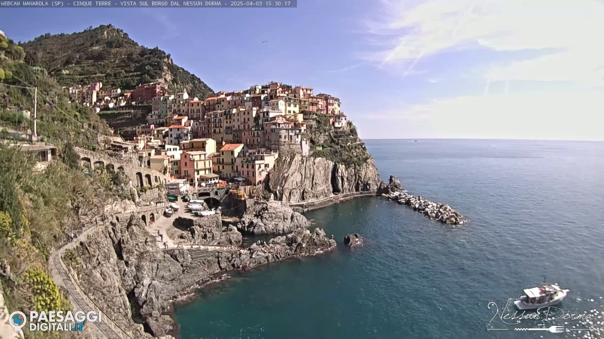 A colorful village clings to a steep cliffside overlooking a calm blue sea under a mostly sunny sky.