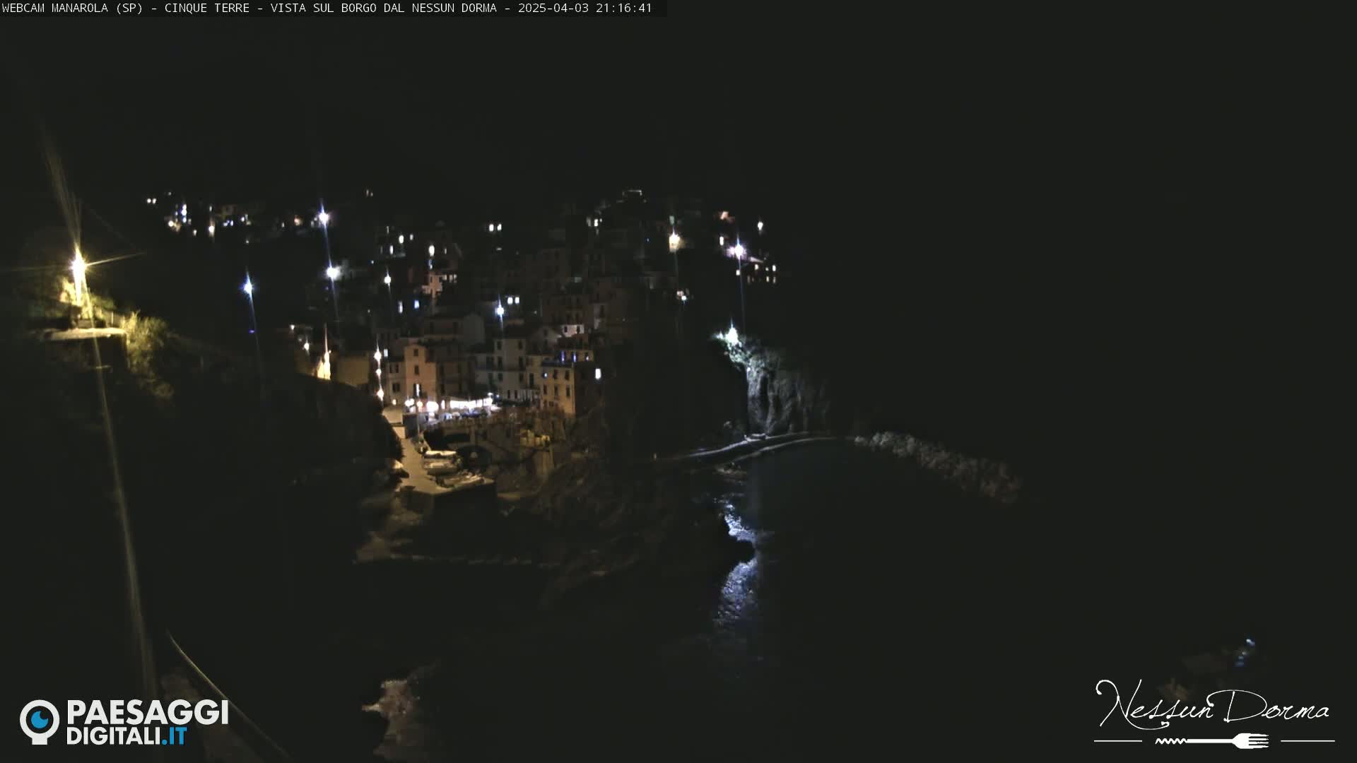 A nighttime, aerial view of a coastal village with buildings illuminated along a dark cliffside overlooking a calm body of water.