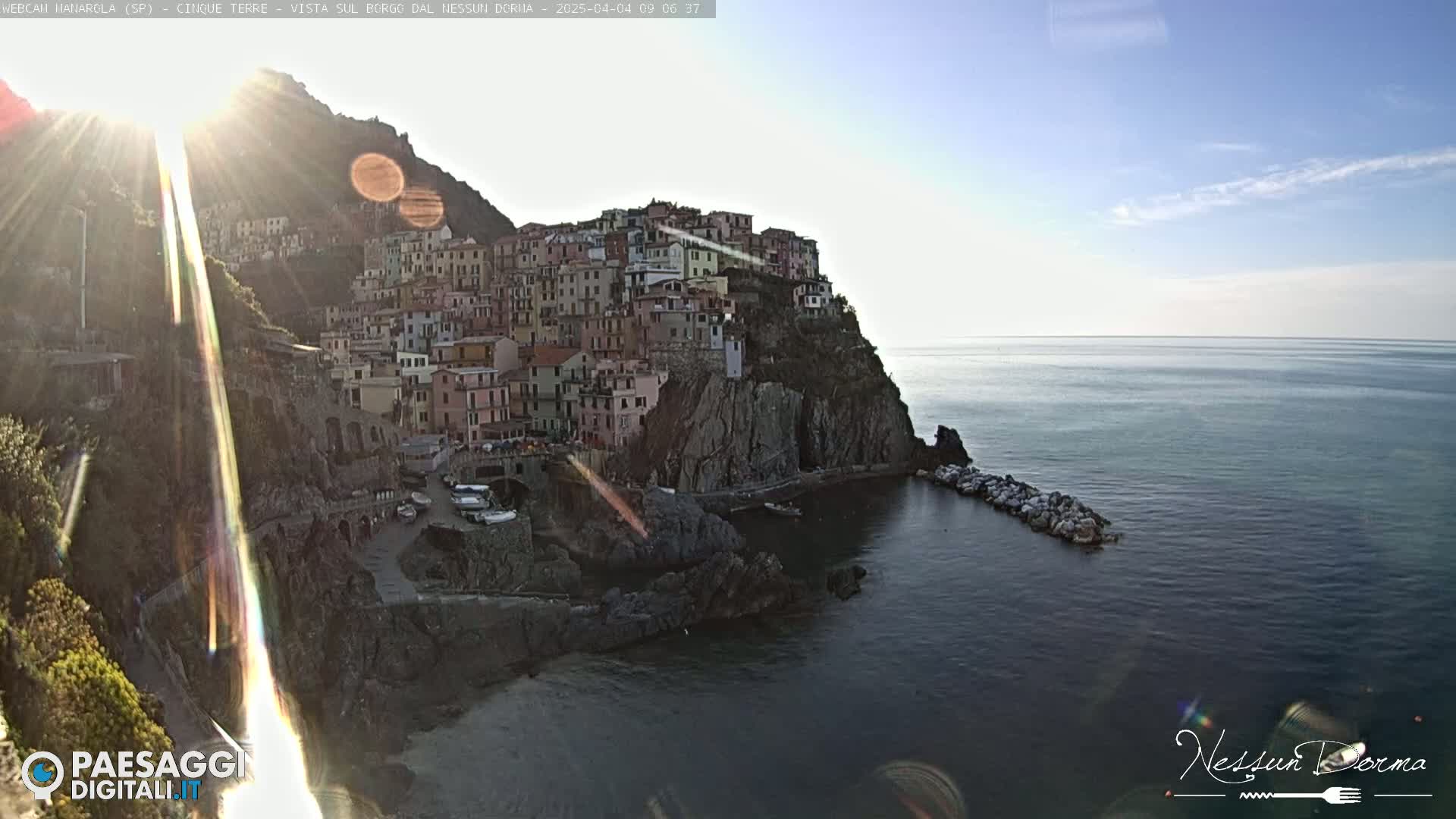A sunlit coastal town, built into a rocky cliffside, overlooks a calm sea on a bright, clear day.