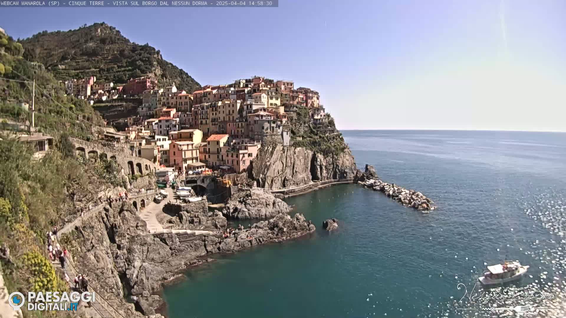A colorful village clings to a rocky cliffside overlooking a calm, blue sea on a sunny day.