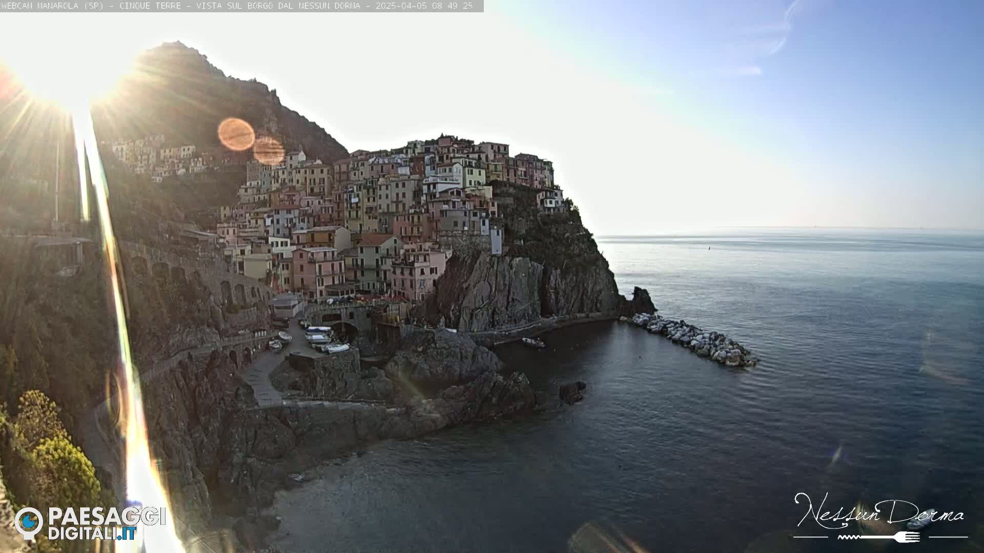 A sunlit coastal village clings to a steep cliffside overlooking a calm sea under a clear sky.