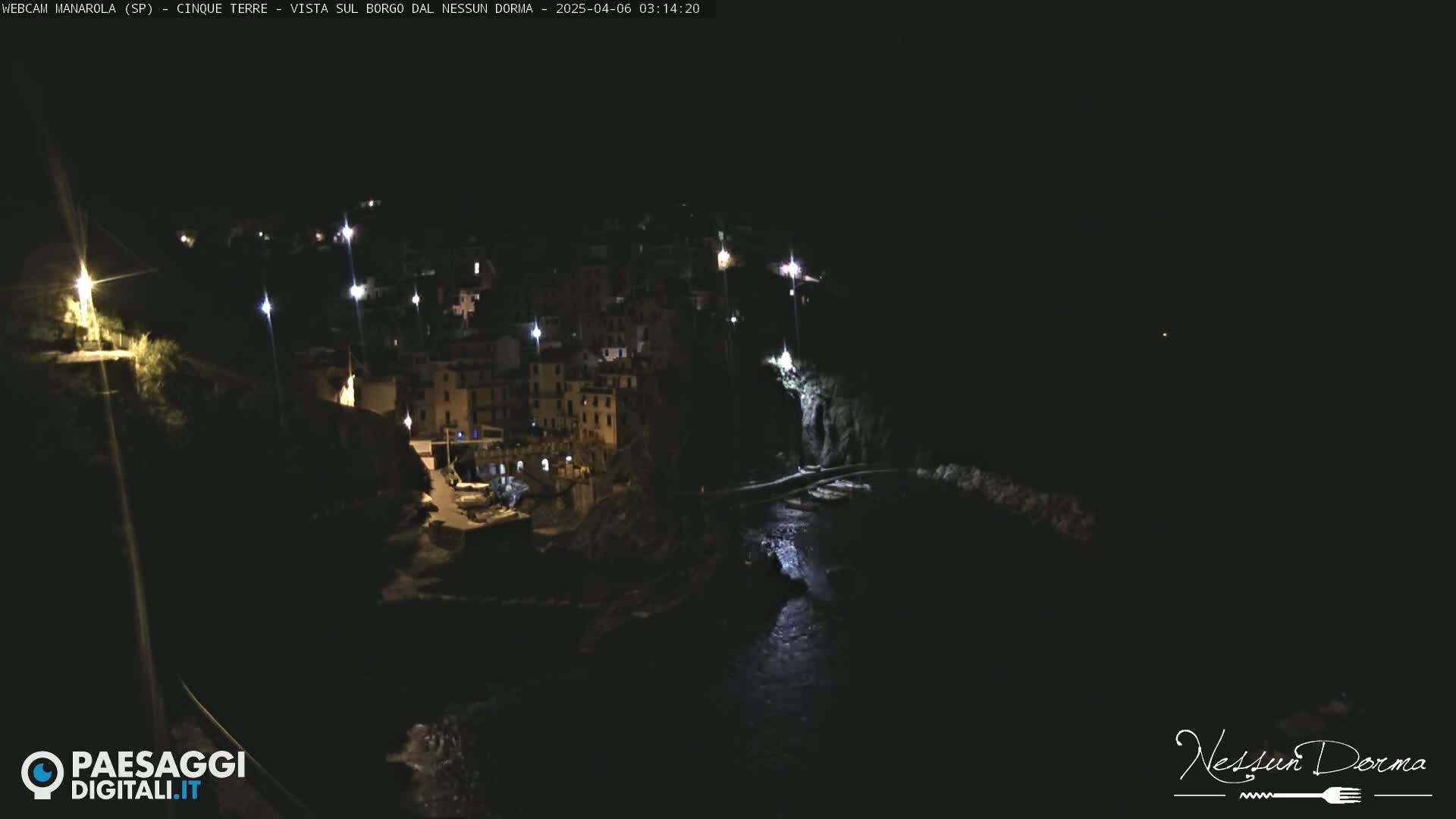 A nighttime, aerial view of a coastal town nestled between dark cliffs, with scattered lights illuminating the buildings and shoreline.
