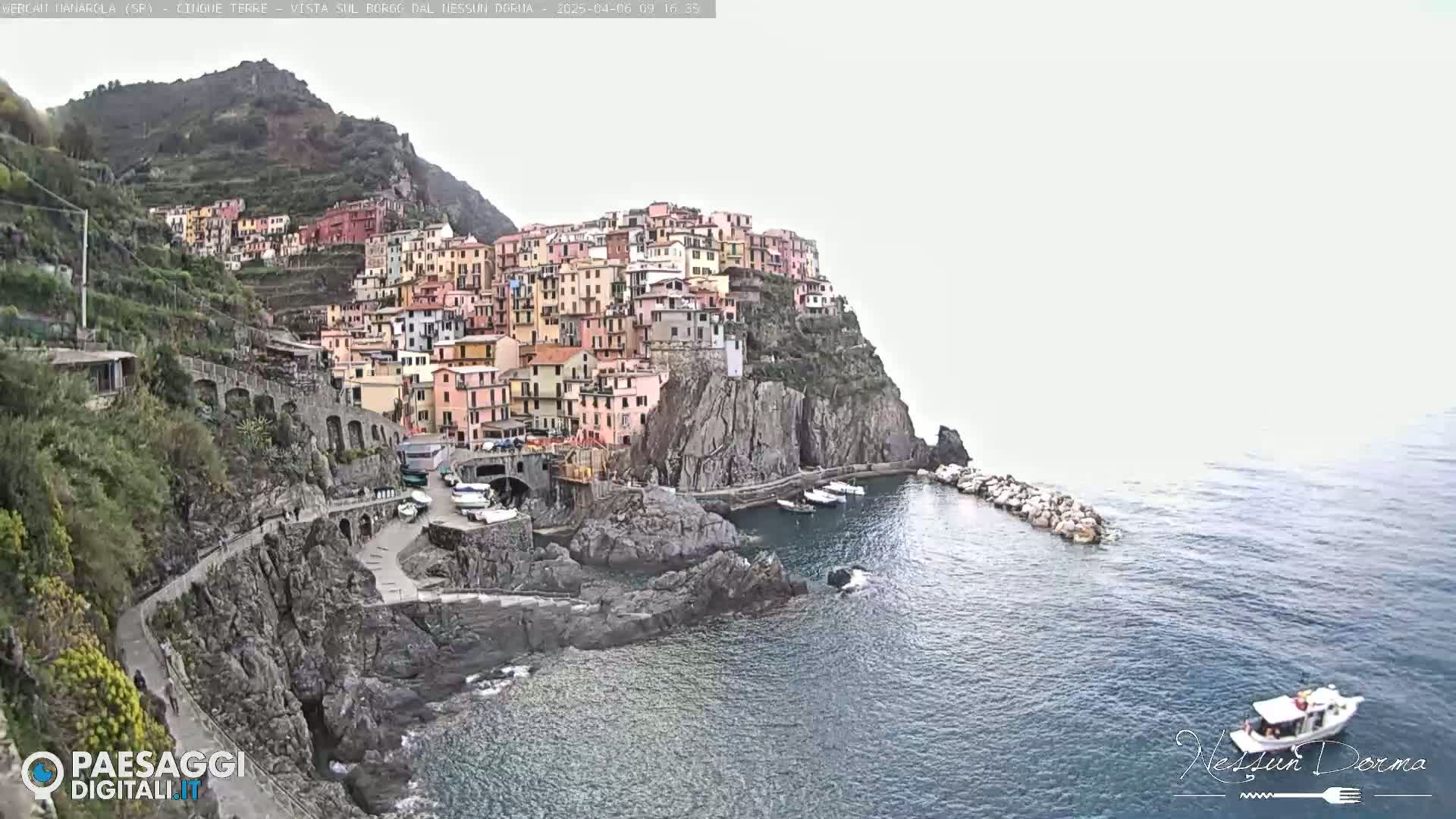 A colorful village clings to a steep cliffside overlooking a calm sea under an overcast sky.