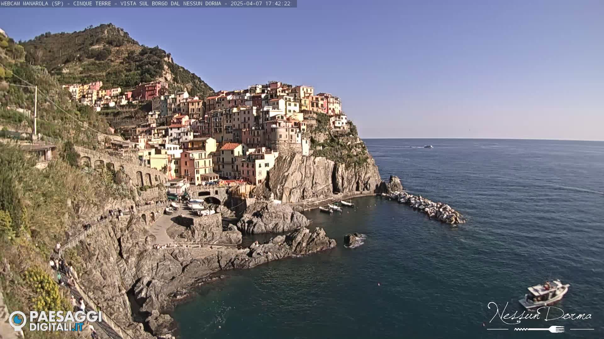 A colorful village clings to a rocky cliffside overlooking a calm, blue sea on a sunny day.